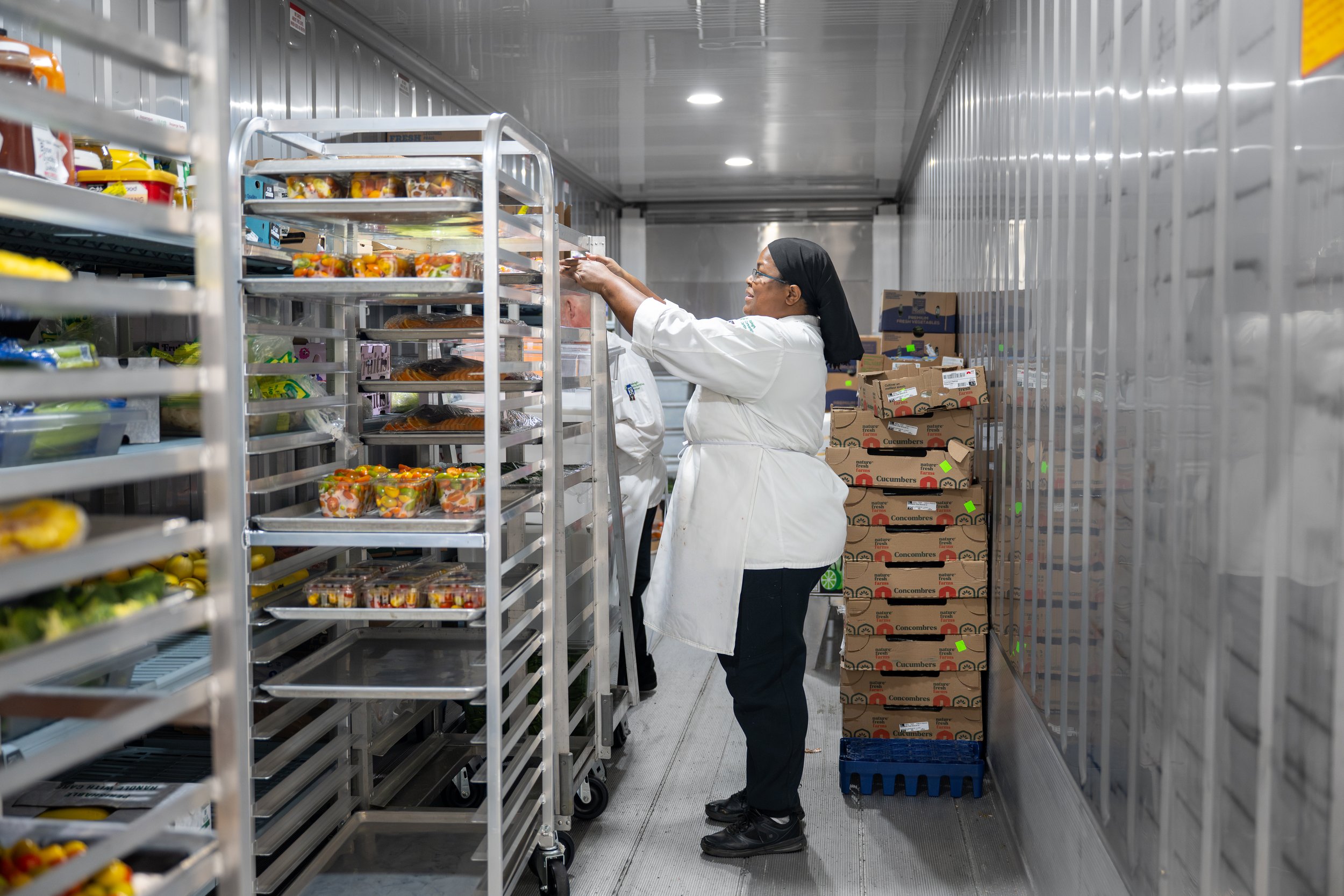 A woman wearing a white chef's coat, glasses, and a black headscarf is stocking pre-packaged fruit salads on a metal shelf in a refrigerated storage room with labeled cardboard boxes of cucumbers and other produce in the background.