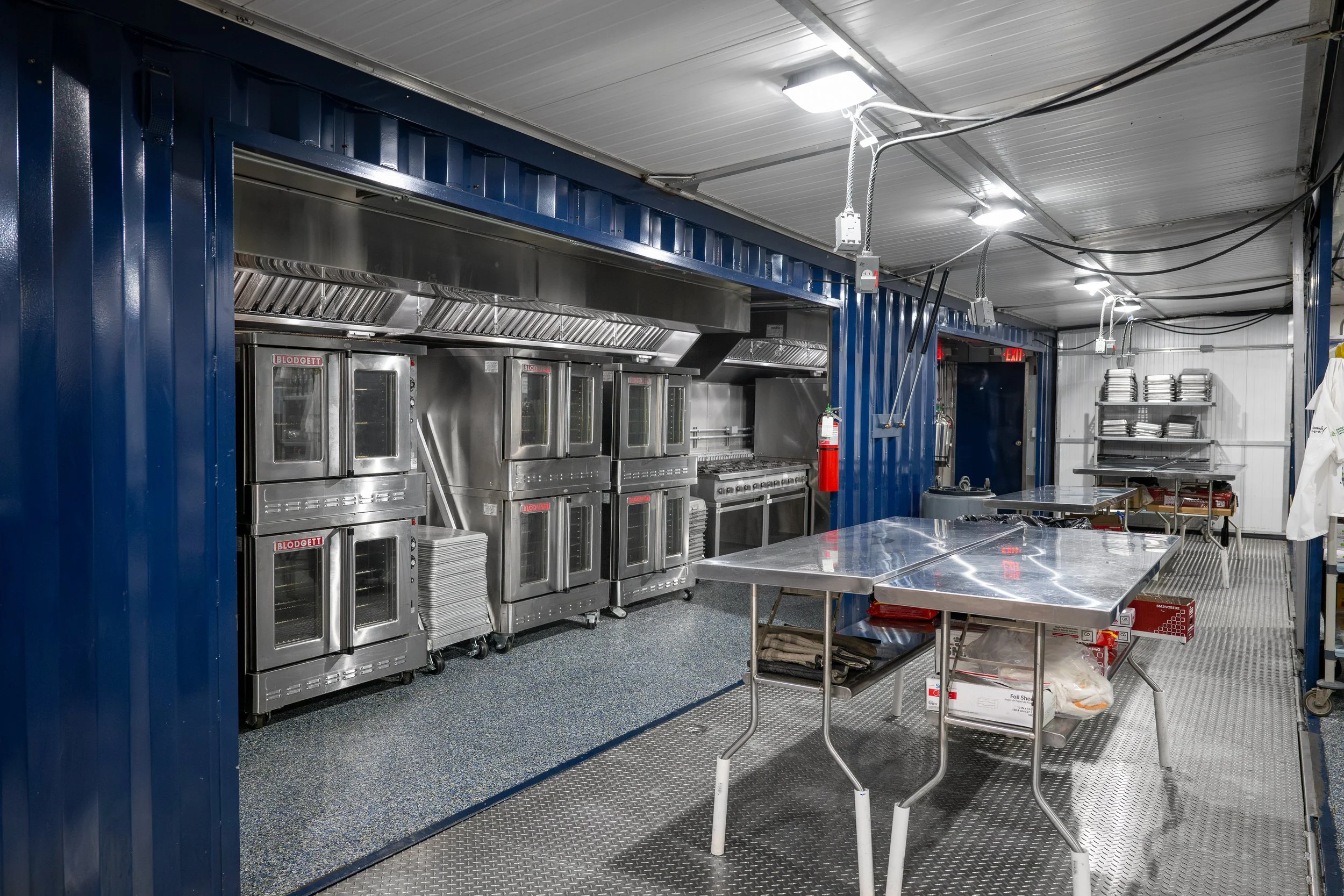 Temporary commercial kitchen with stainless steel appliances, blue walls, metal tables, and shelves with stacked plates and kitchen supplies.