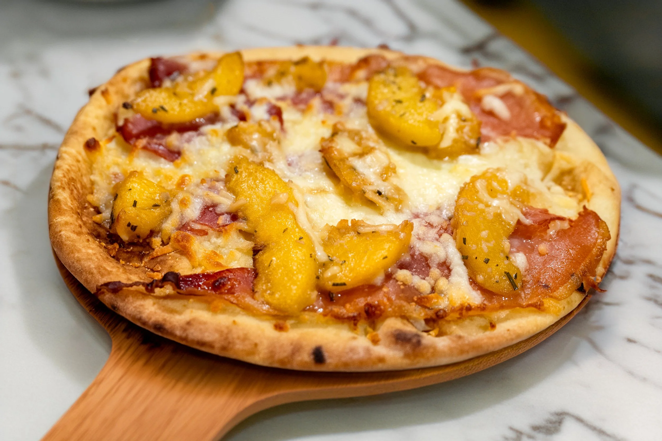 Close-up of a pizza with sliced peaches, ham, and cheese on a wooden paddle.