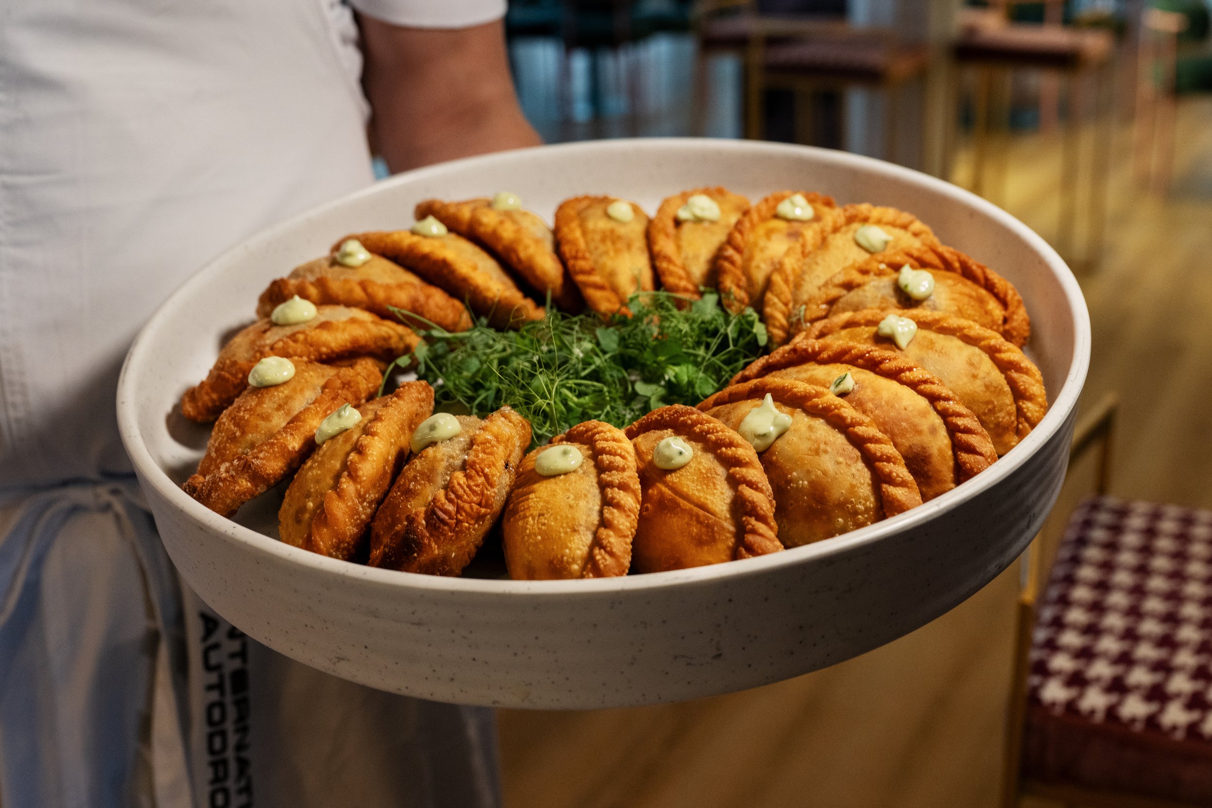 A person holding a bowl of fried empanadas garnished with green herbs and small dollops of white sauce.