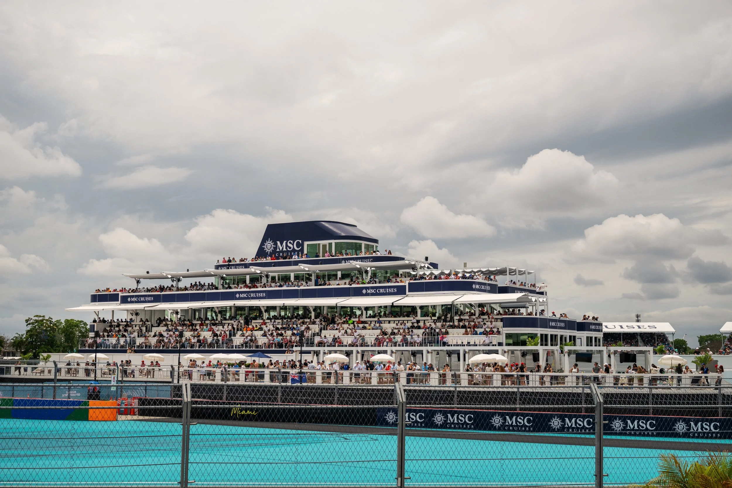 A large MSC Cruises cruise ship with multiple decks filled with people, docked outdoors on a cloudy day, with a swimming pool in the foreground.