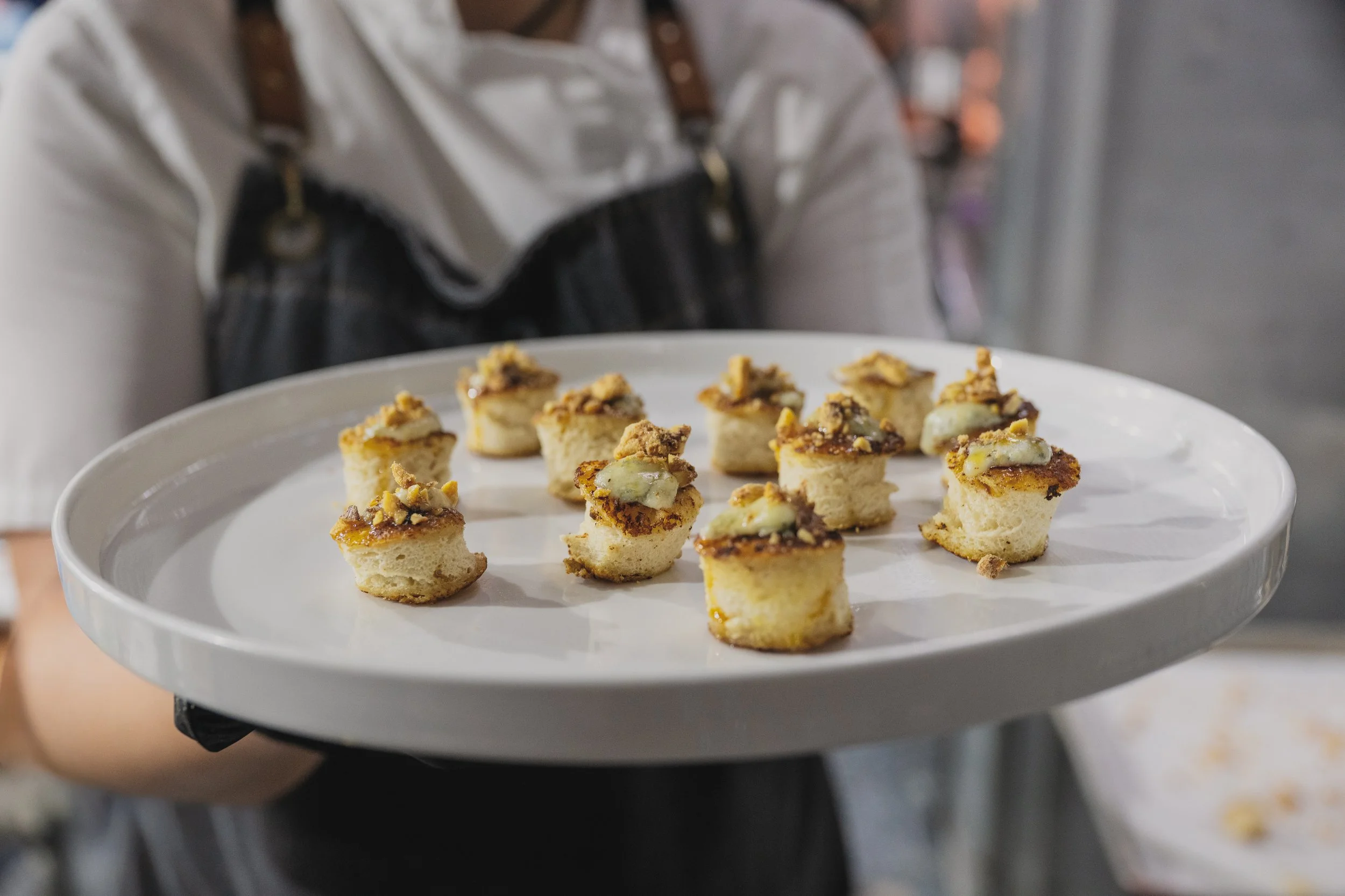A person holding a white plate with small, cannoli-like desserts topped with nuts and white icing. The background is blurred.
