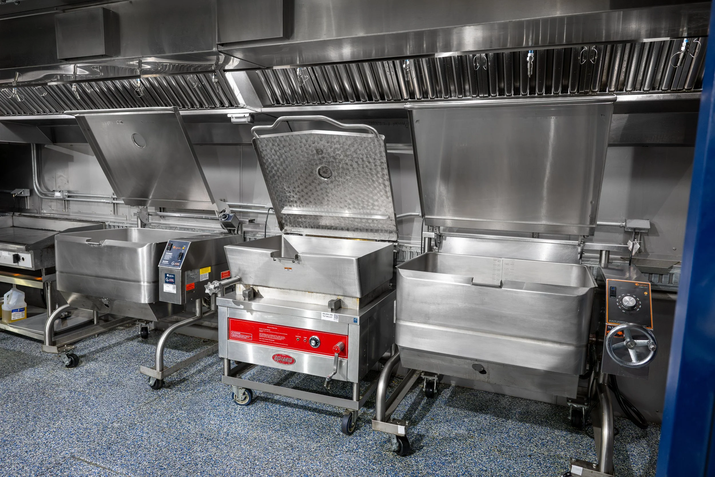 A row of stainless steel industrial kitchen equipment, including large tilt skillets, in a commercial kitchen setting.