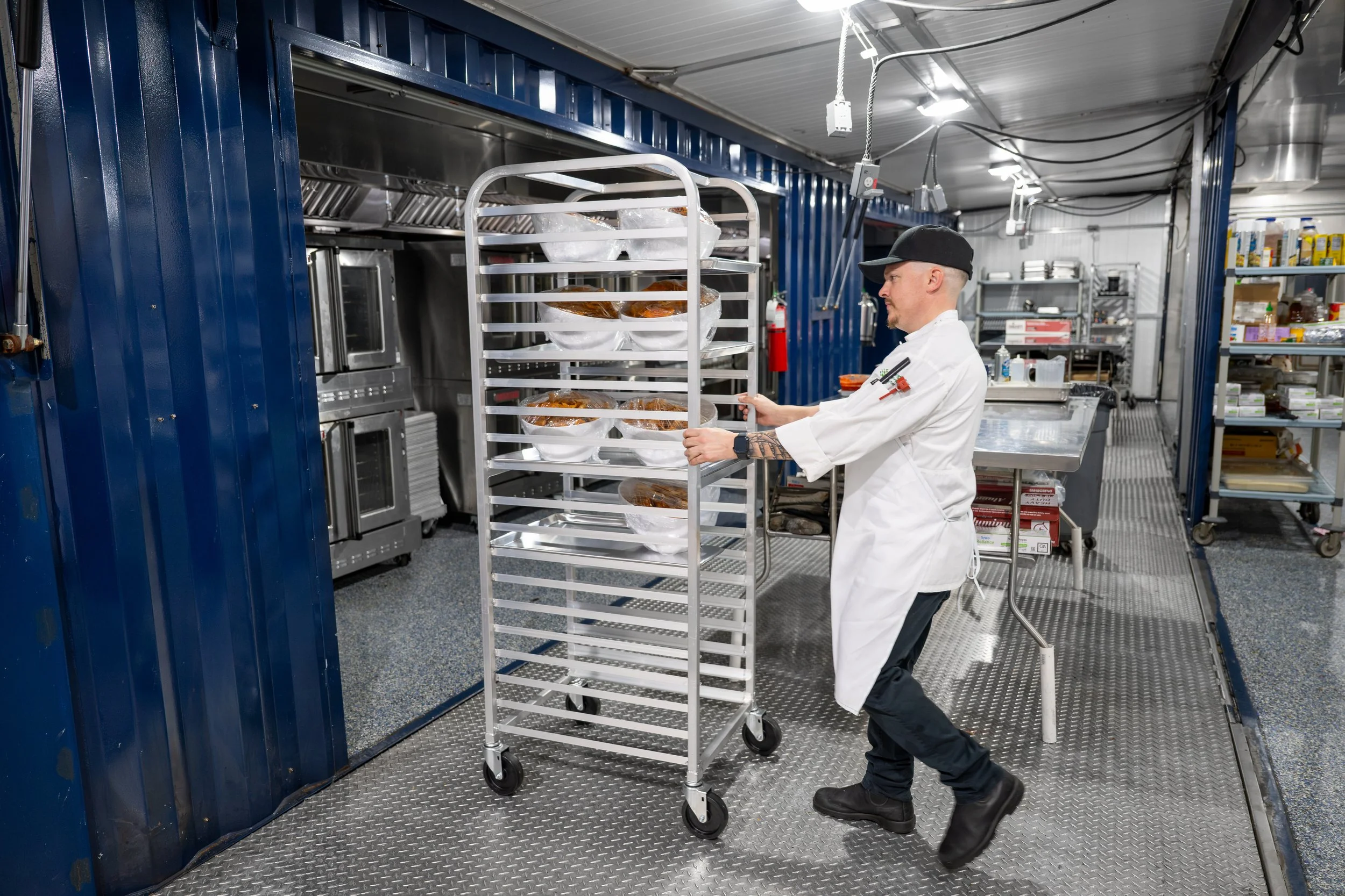 A chef in a white uniform and black cap pushes a metal rack with plastic-wrapped food packages in a commercial kitchen with metal shelves, appliances, and a blue wall.
