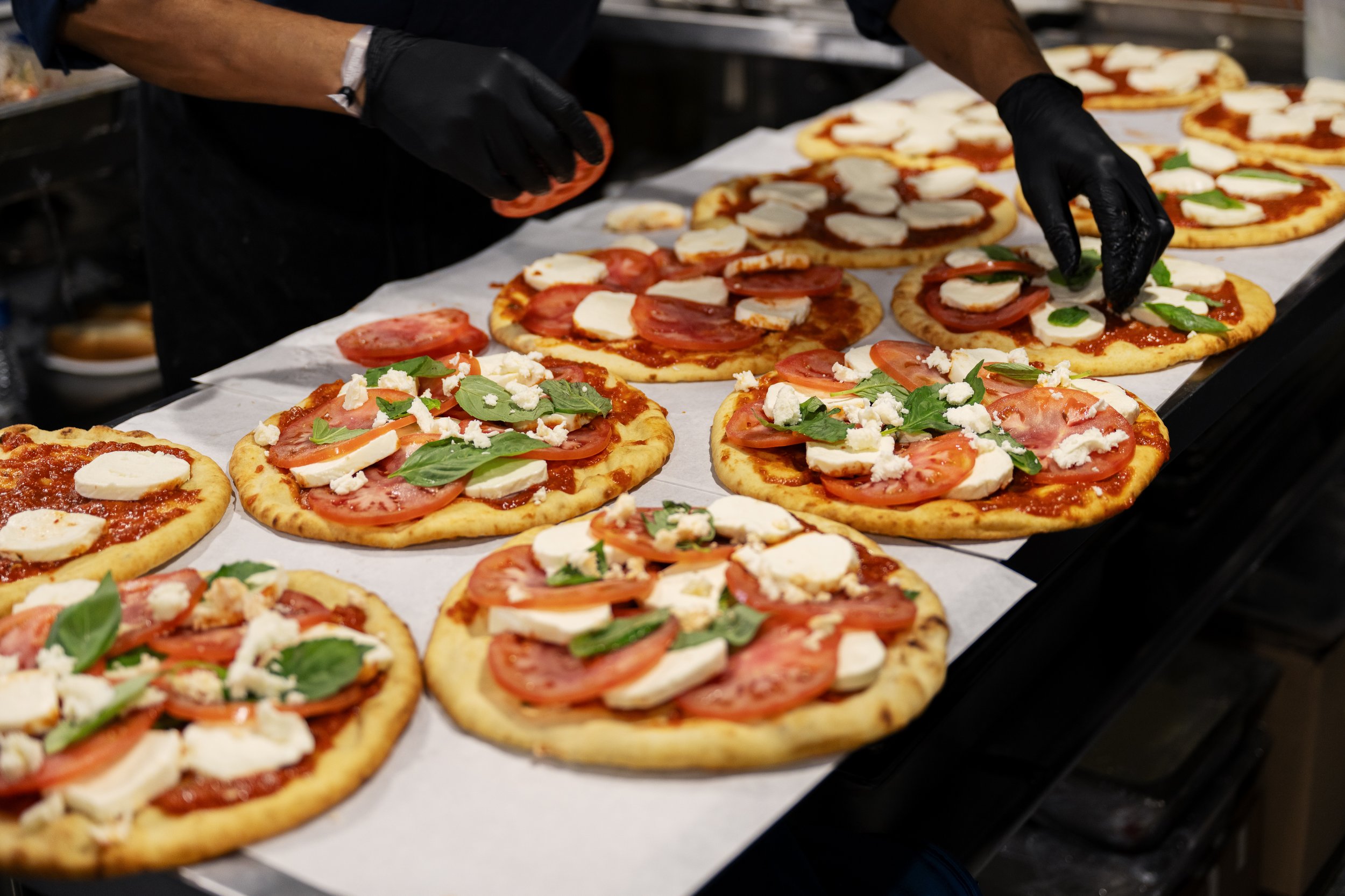 Person in black gloves arranging pizzas topped with tomatoes, mozzarella, basil, and other ingredients on a tray.