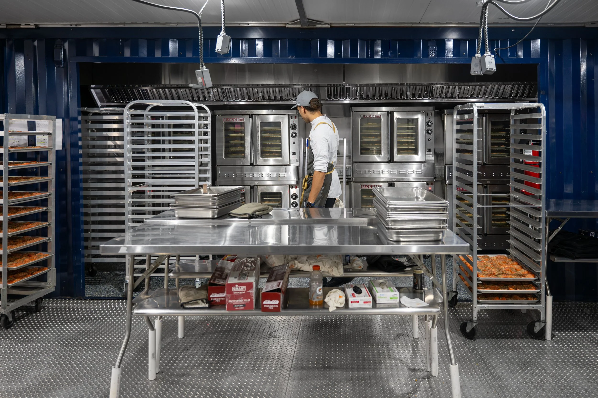 Commercial kitchen with a person inspecting bakers in front of ovens, surrounded by racks of trays of food and baking supplies.