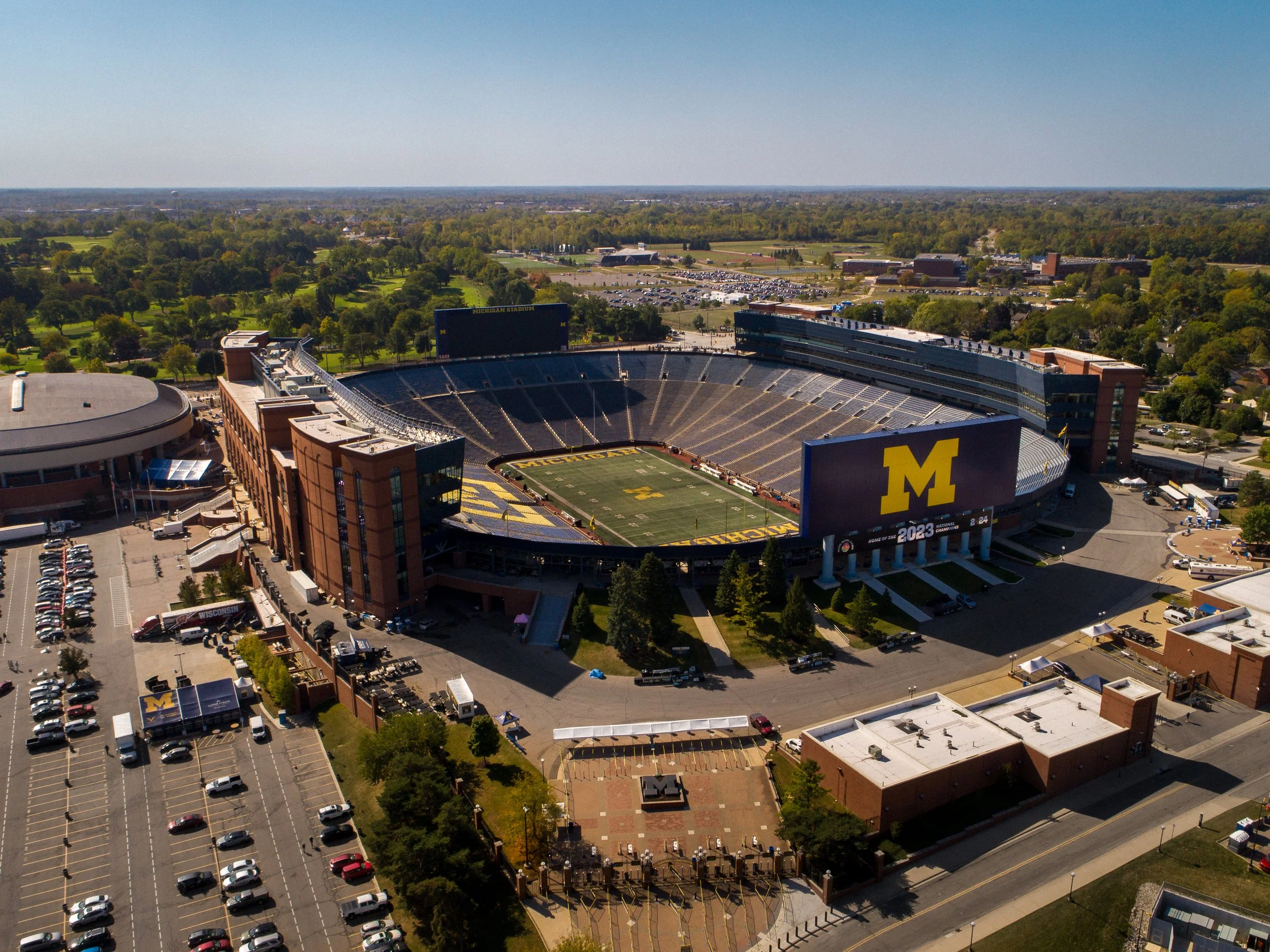 Aerial view of Michigan Stadium in Ann Arbor, Michigan, with the football field in the center surrounded by seating, parking lot, and trees.