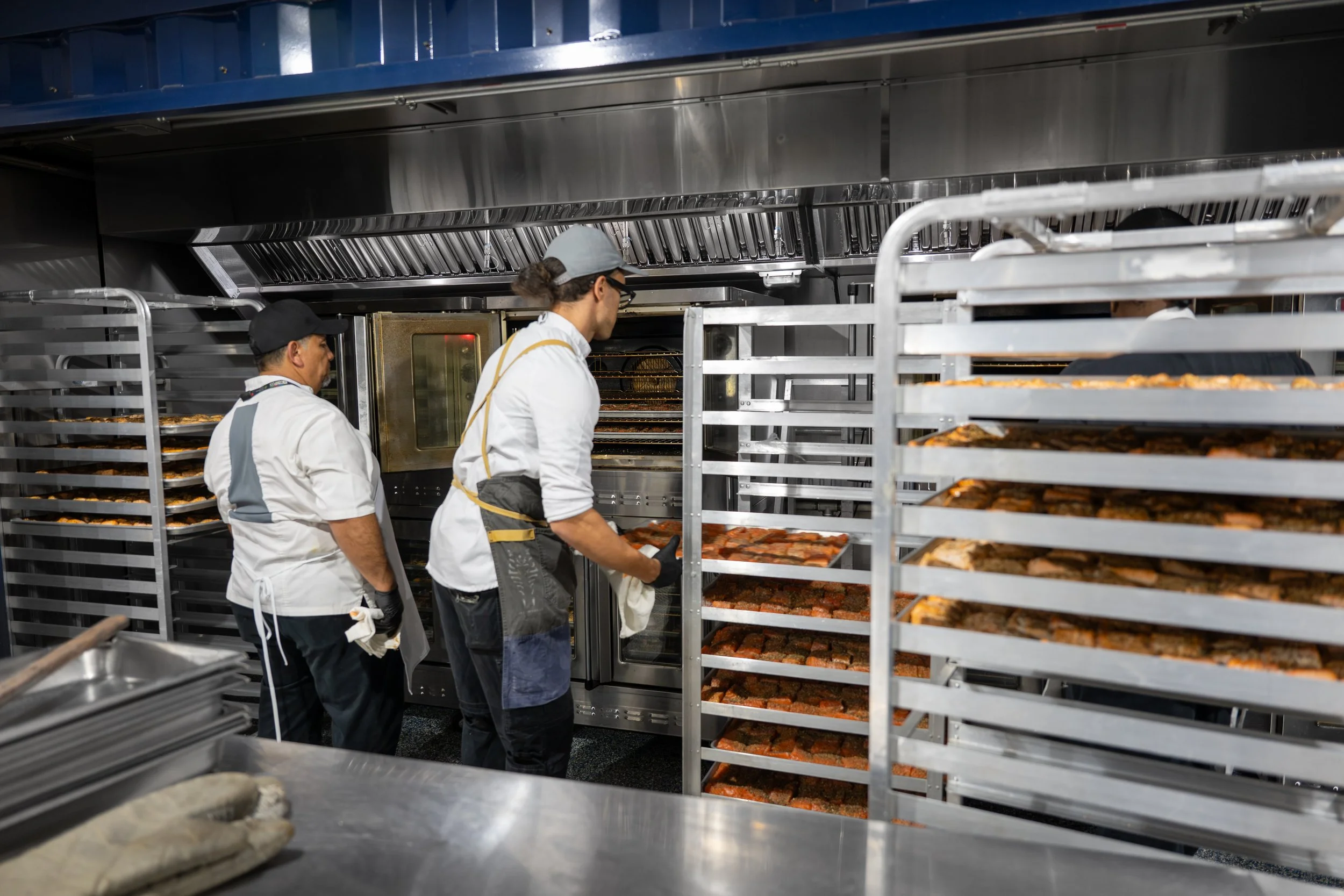 People working in a commercial kitchen preparing food, with multiple racks of baked or cooked items and kitchen equipment.
