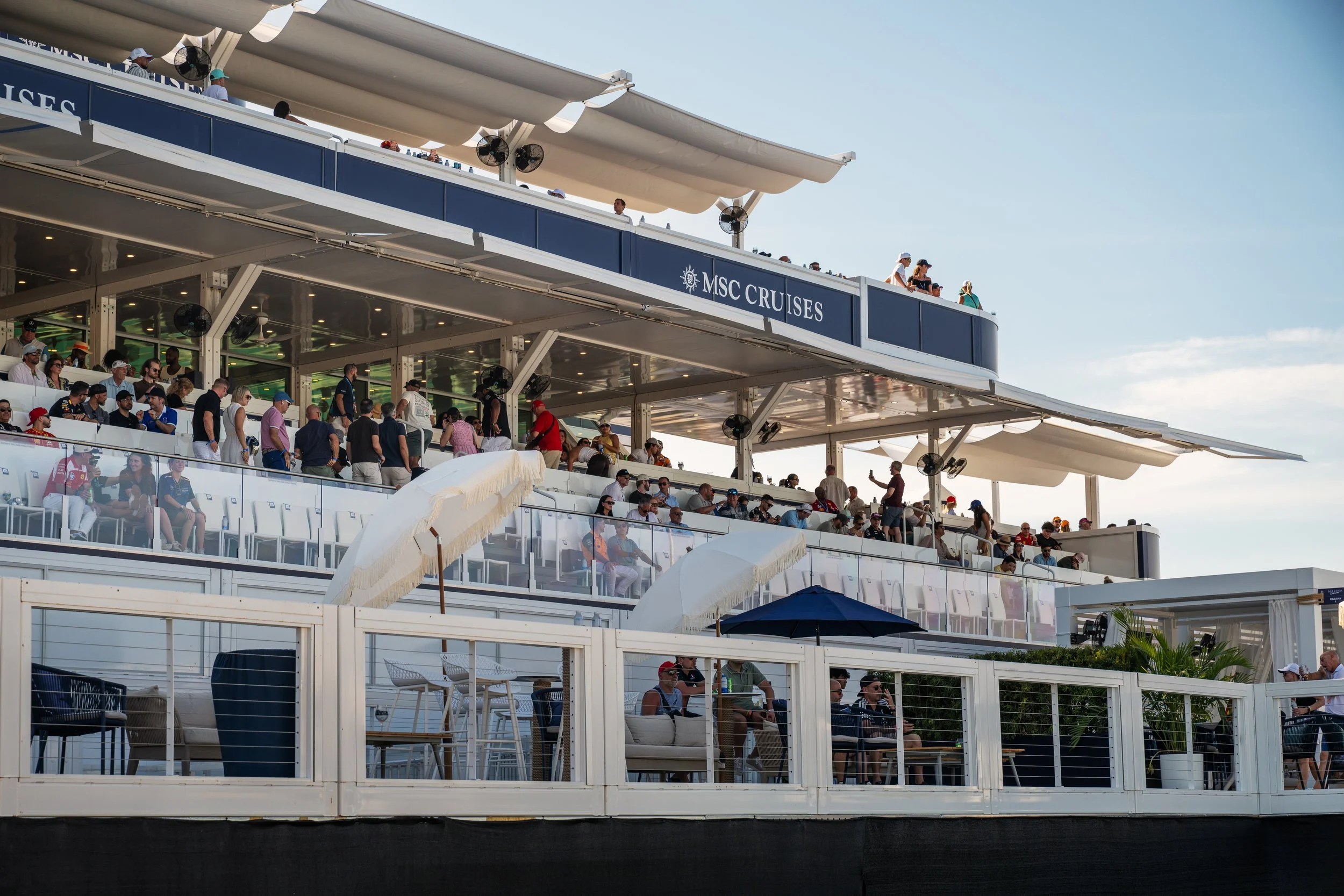 Multiple levels of a cruise ship's deck filled with people seated and standing, some watching an event or view, with bright blue sky in the background.