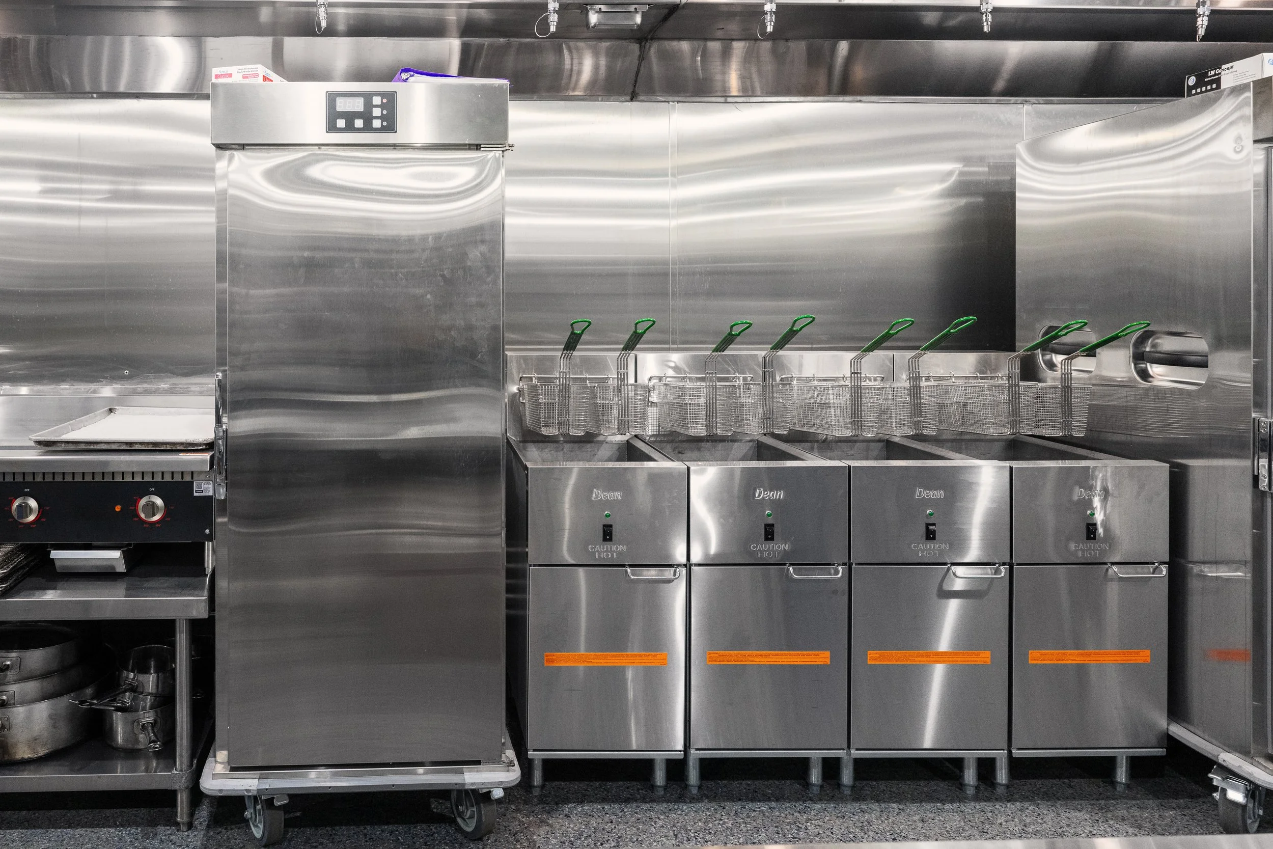 Commercial kitchen equipment with multiple stainless steel appliances, including fryers with green handles on the right, and an oven or warmer in the top left corner, all set against stainless steel walls.