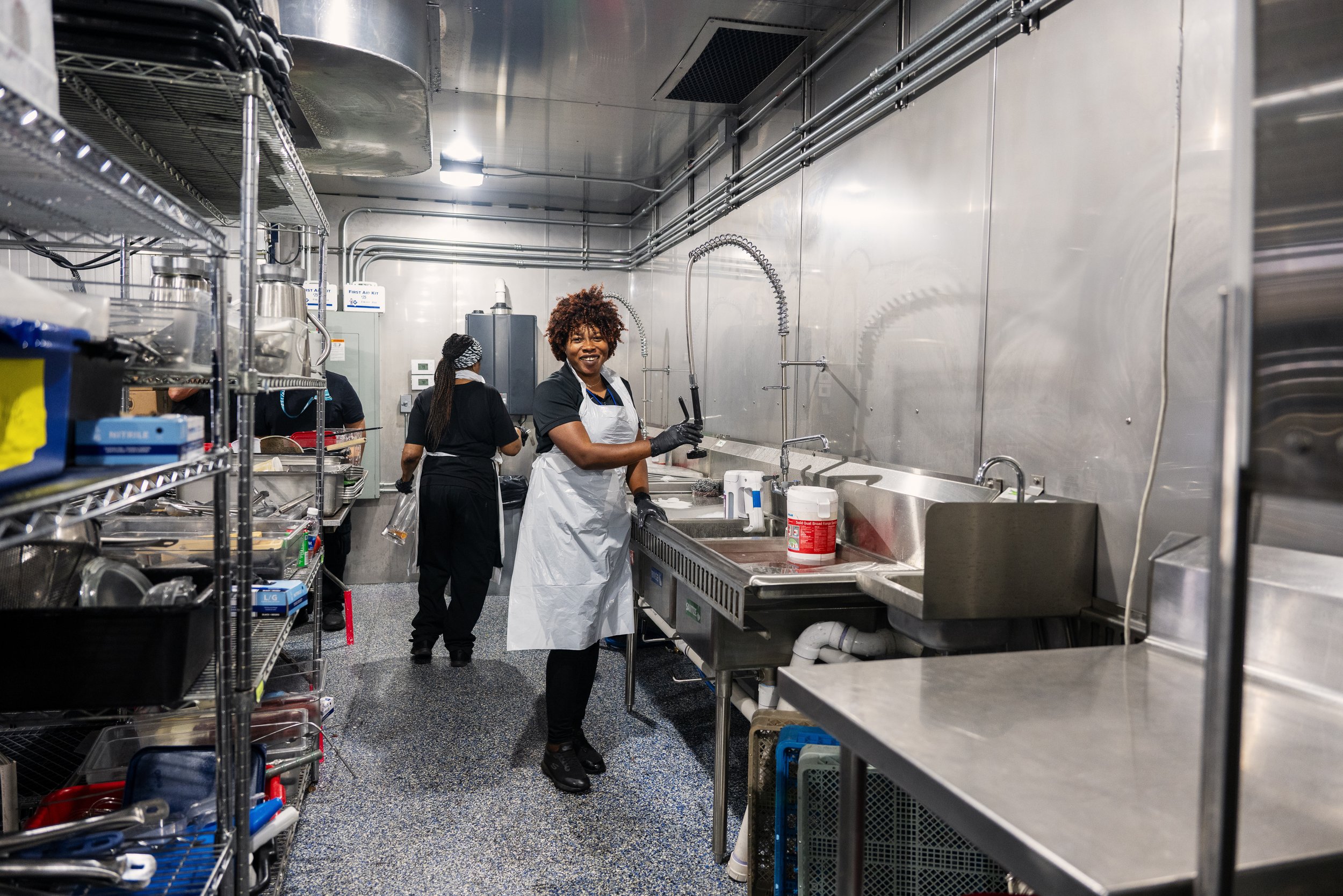 Three women in a commercial kitchen, one in front smiling at the camera wearing a white apron and black gloves, two others in the background working near stainless steel counters and kitchen shelves filled with supplies and utensils.