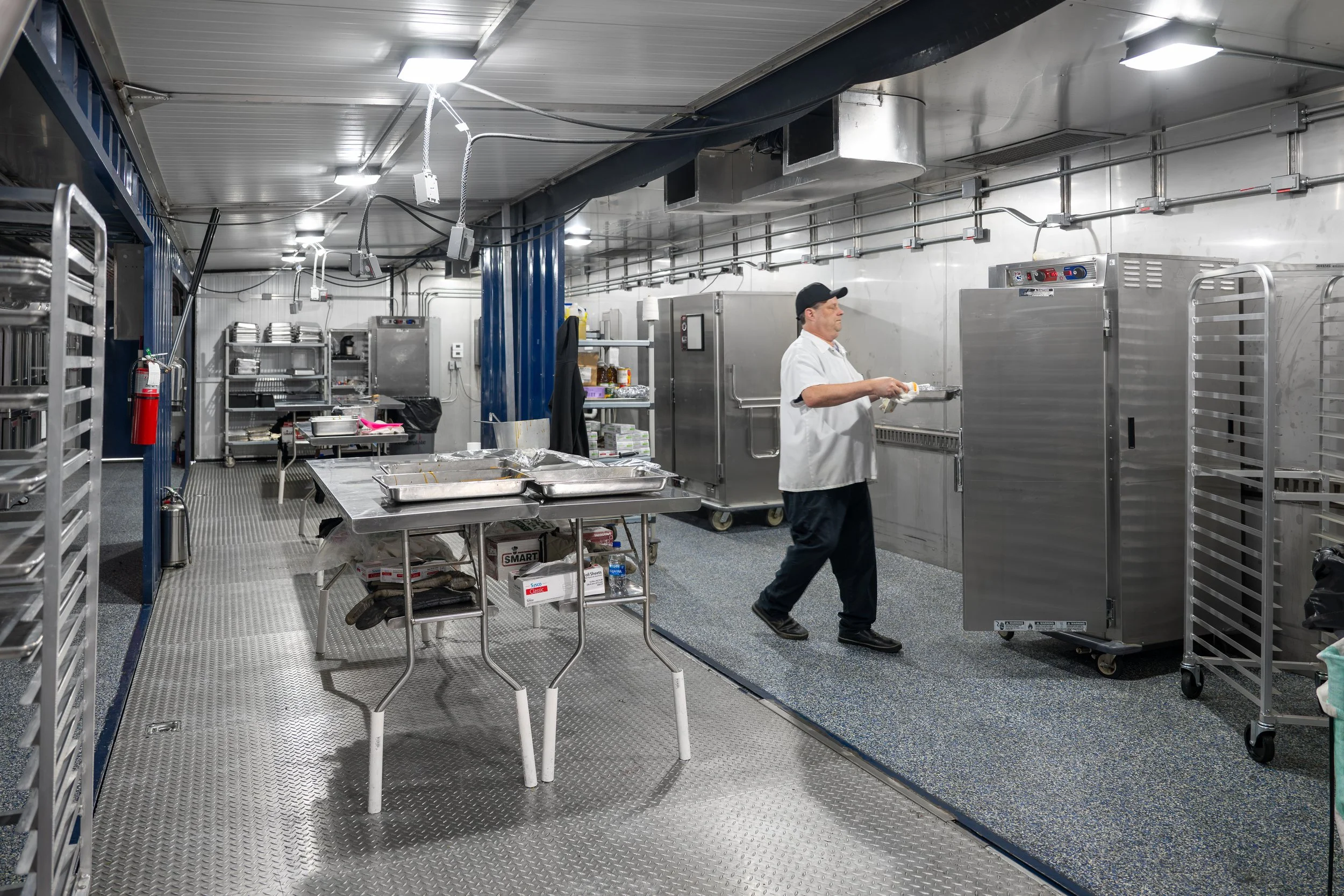A man in a white uniform and black cap is walking in a commercial kitchen or food processing facility, holding a tray. The room contains stainless steel equipment, shelving, carts, and a metal floor, with overhead lights.