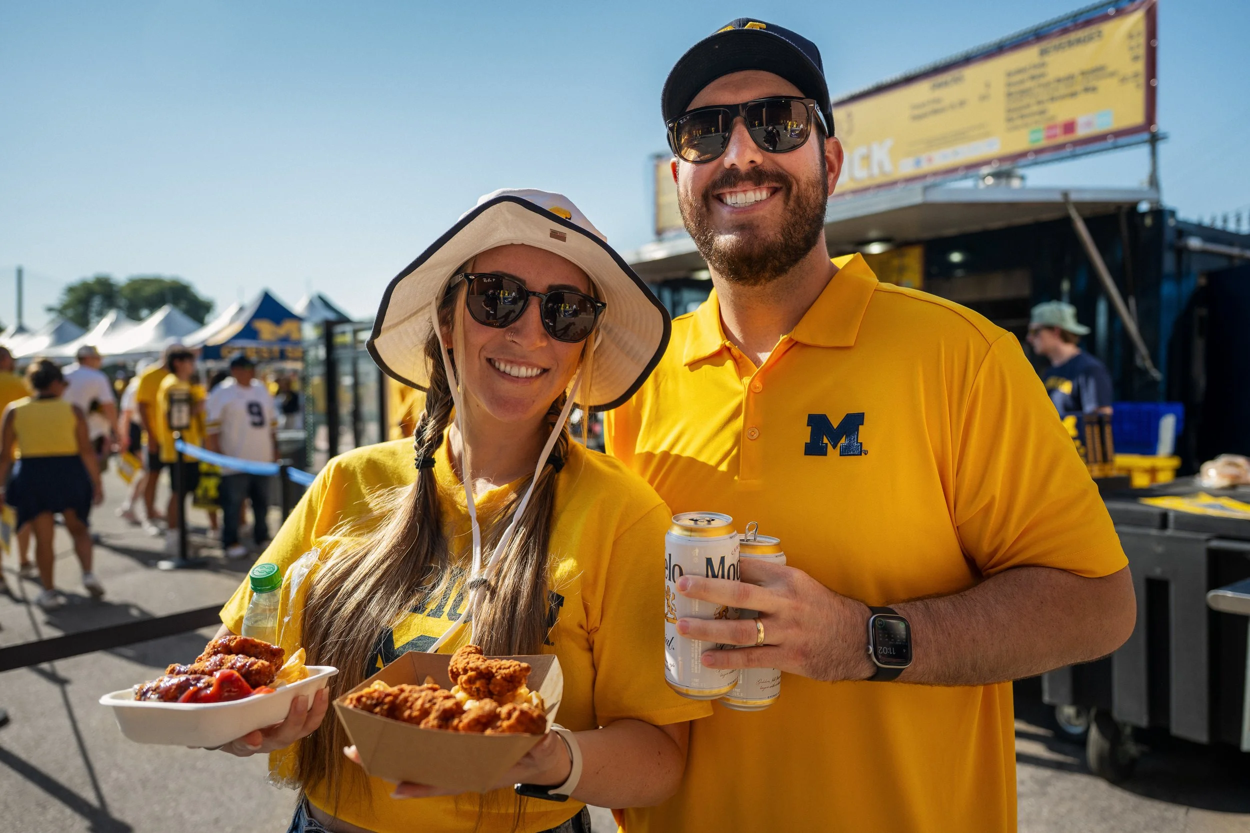 Two friends, a woman with long hair, a hat, and sunglasses, and a man with a beard, sunglasses, and a yellow Michigan Wolverines polo shirt, are smiling and holding food and drinks at an outdoor event on a sunny day. They are surrounded by other peop