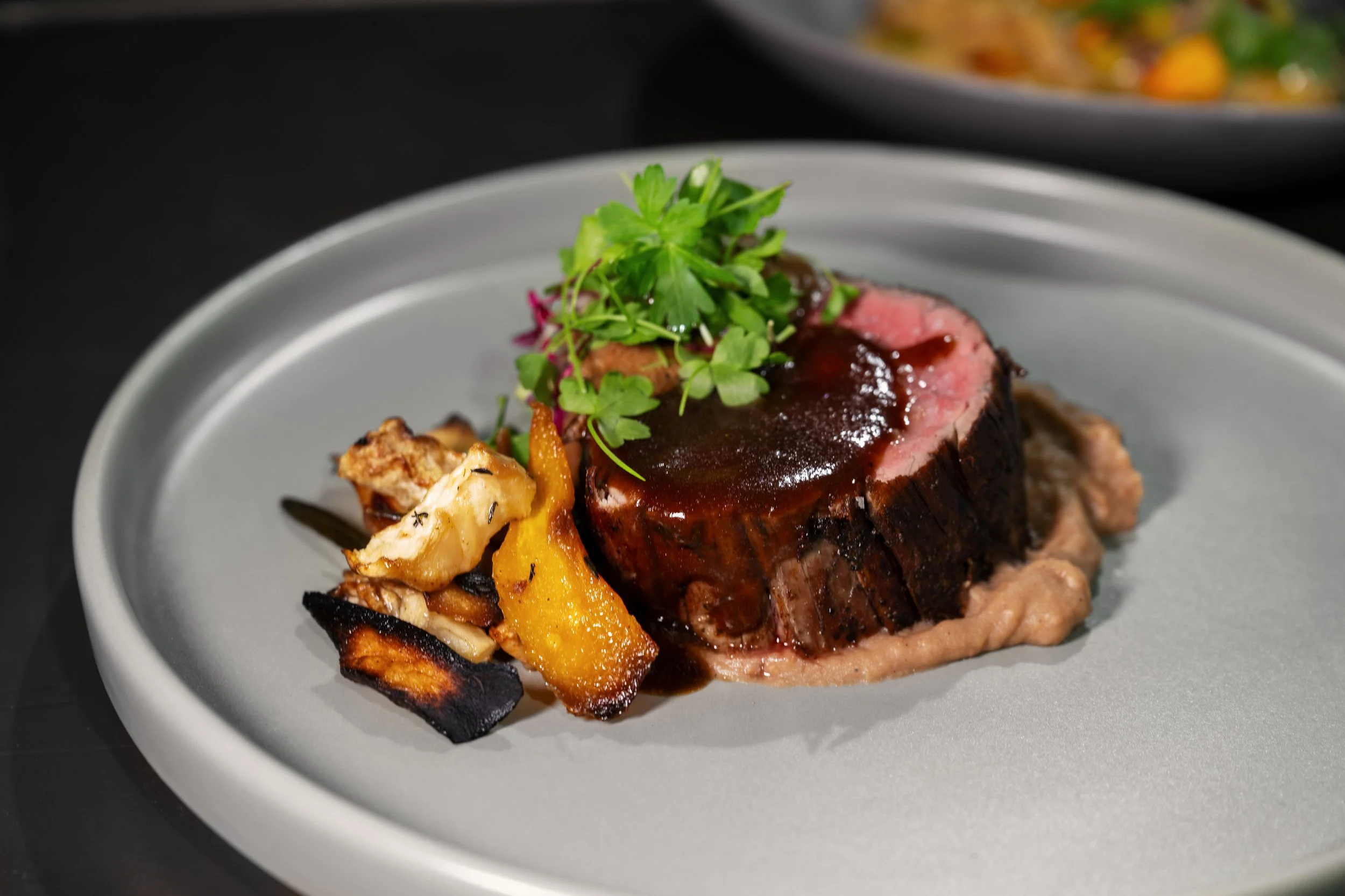 A plate with a medium-rare steak topped with green herbs, accompanied by roasted vegetables, on a gray dish with a black background.