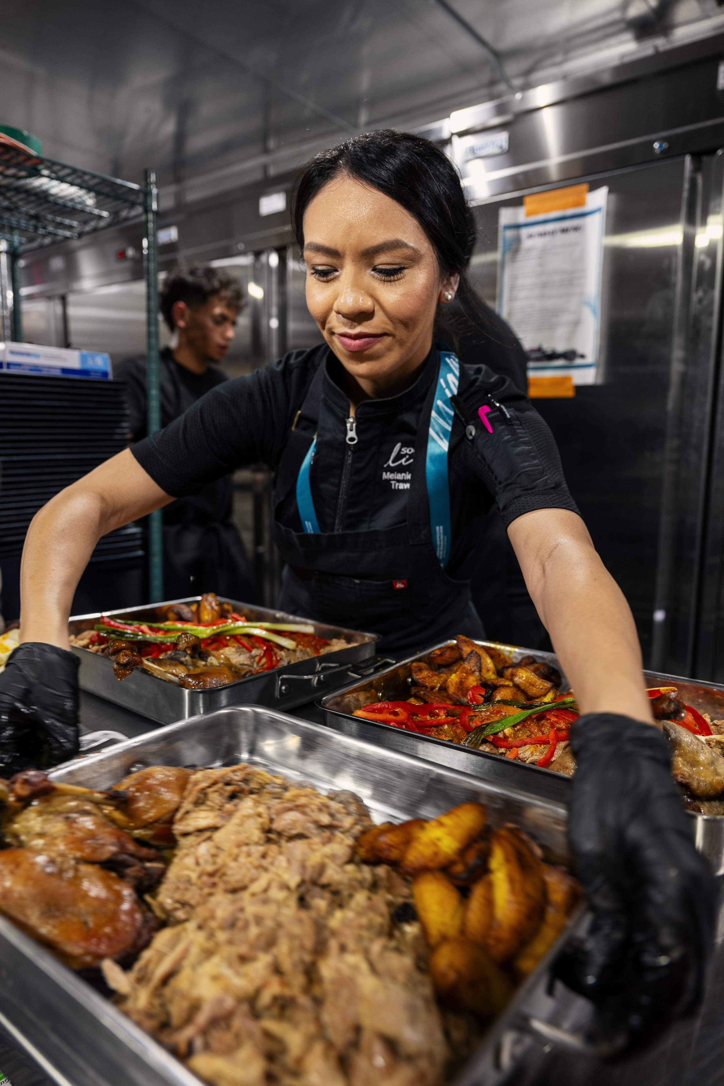 A woman with black hair, wearing black gloves, is serving herself food from multiple trays of cooked dishes, including meats and vegetables, in a commercial kitchen or catering setting. Behind her, a young man is working with food, and there are shel