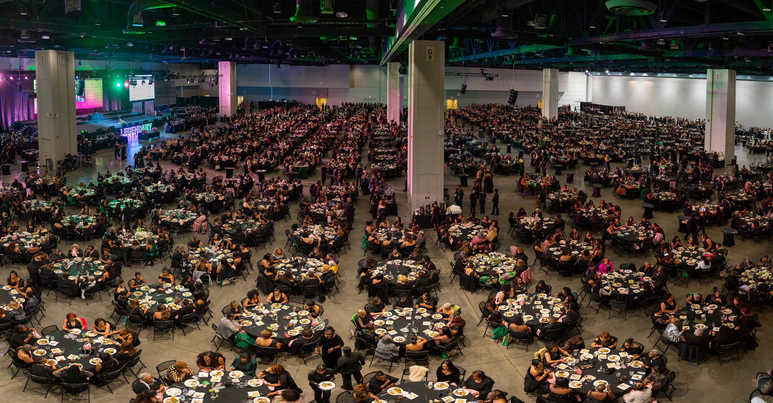 A large indoor event with many round tables set for a meal, filled with people, under colorful stage lighting in a convention hall.
