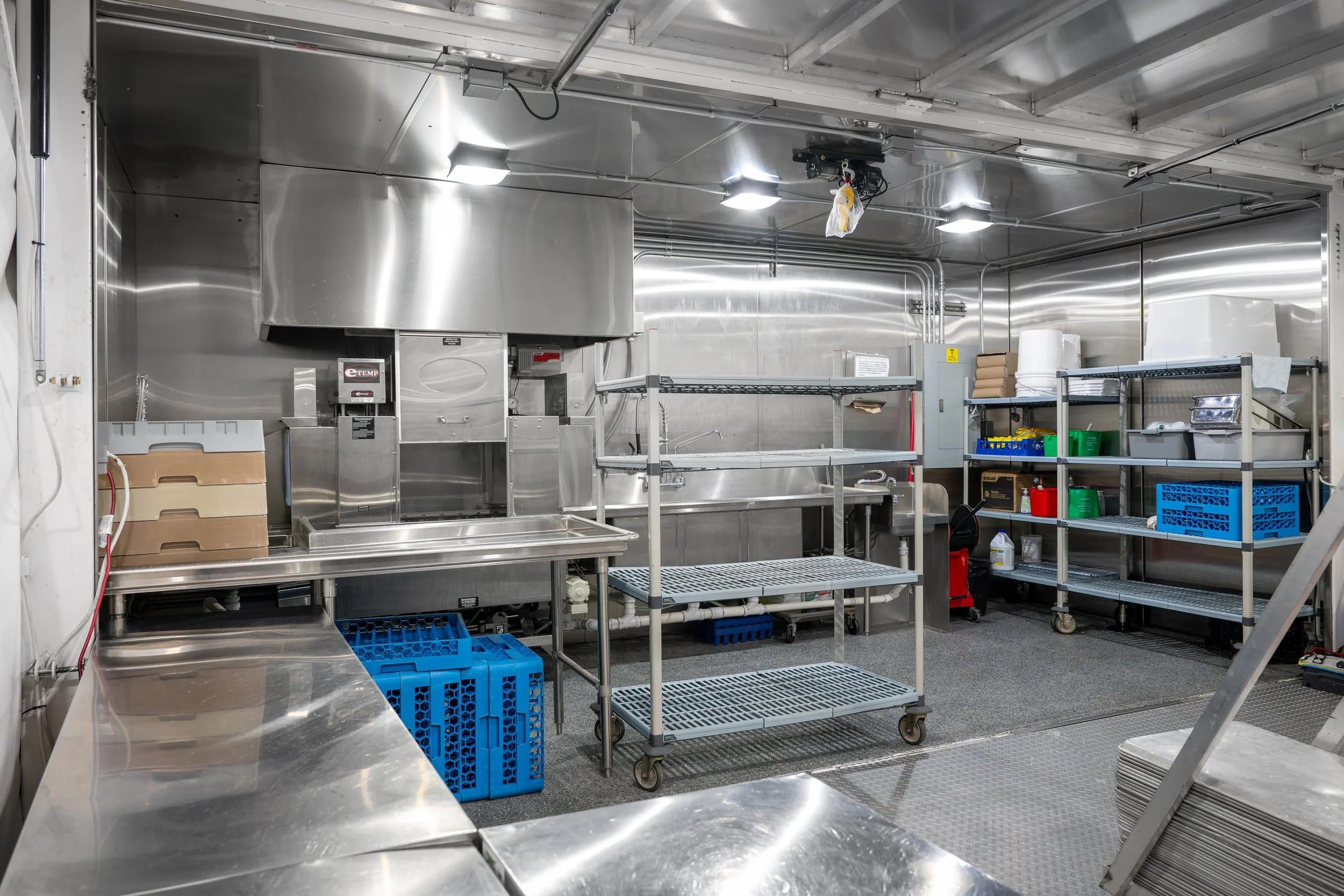 A commercial kitchen storage area with stainless steel work surfaces, shelves, and equipment. Contains plastic crates, boxes, and rolls of paper or foil.