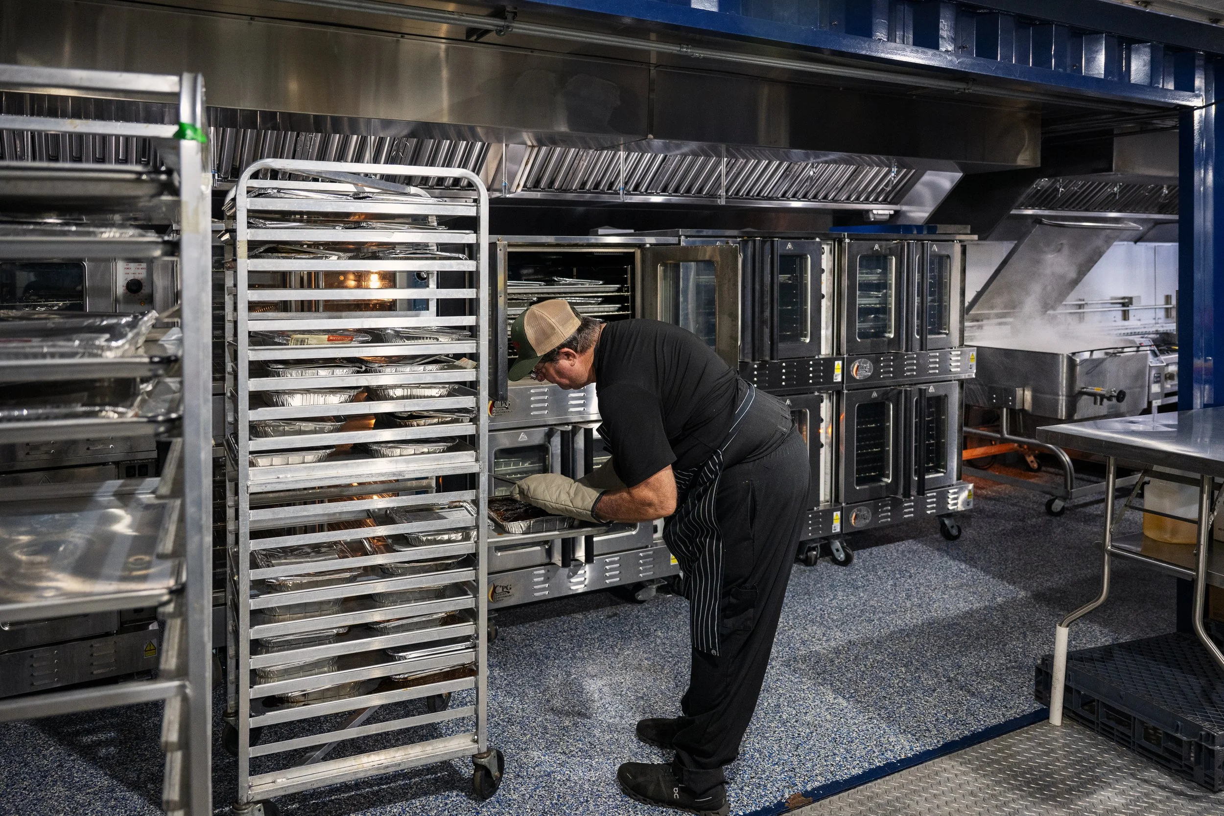 A chef wearing a baseball cap, black t-shirt, and striped apron working in a commercial kitchen. He is leaning over a tray, tending to food items. Surrounding him are multiple stainless steel racks and industrial ovens.