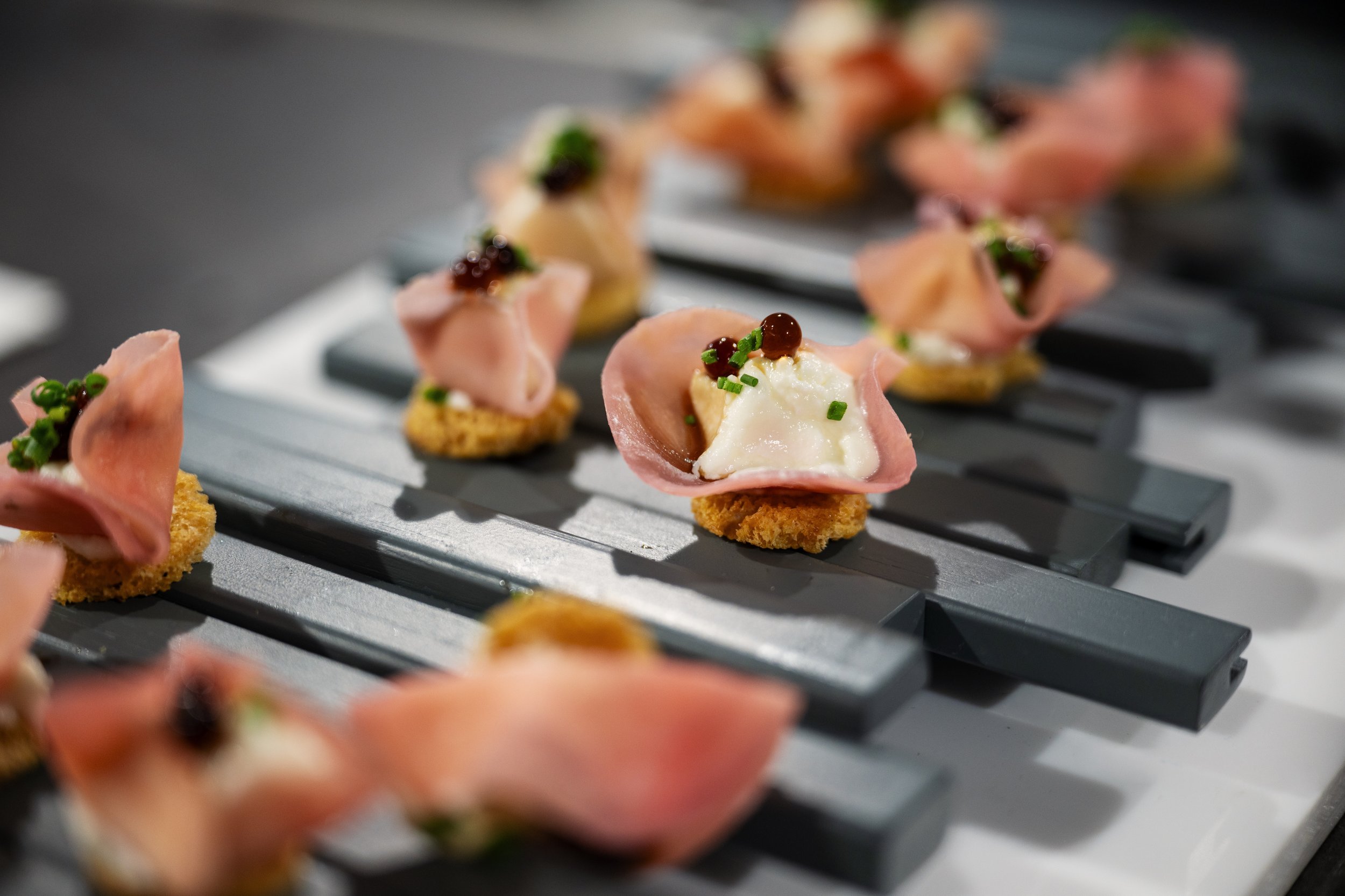 Close-up of canapé appetizers with sliced meat, cheese, garnished with herbs and small pearls, arranged on a gray serving tray.