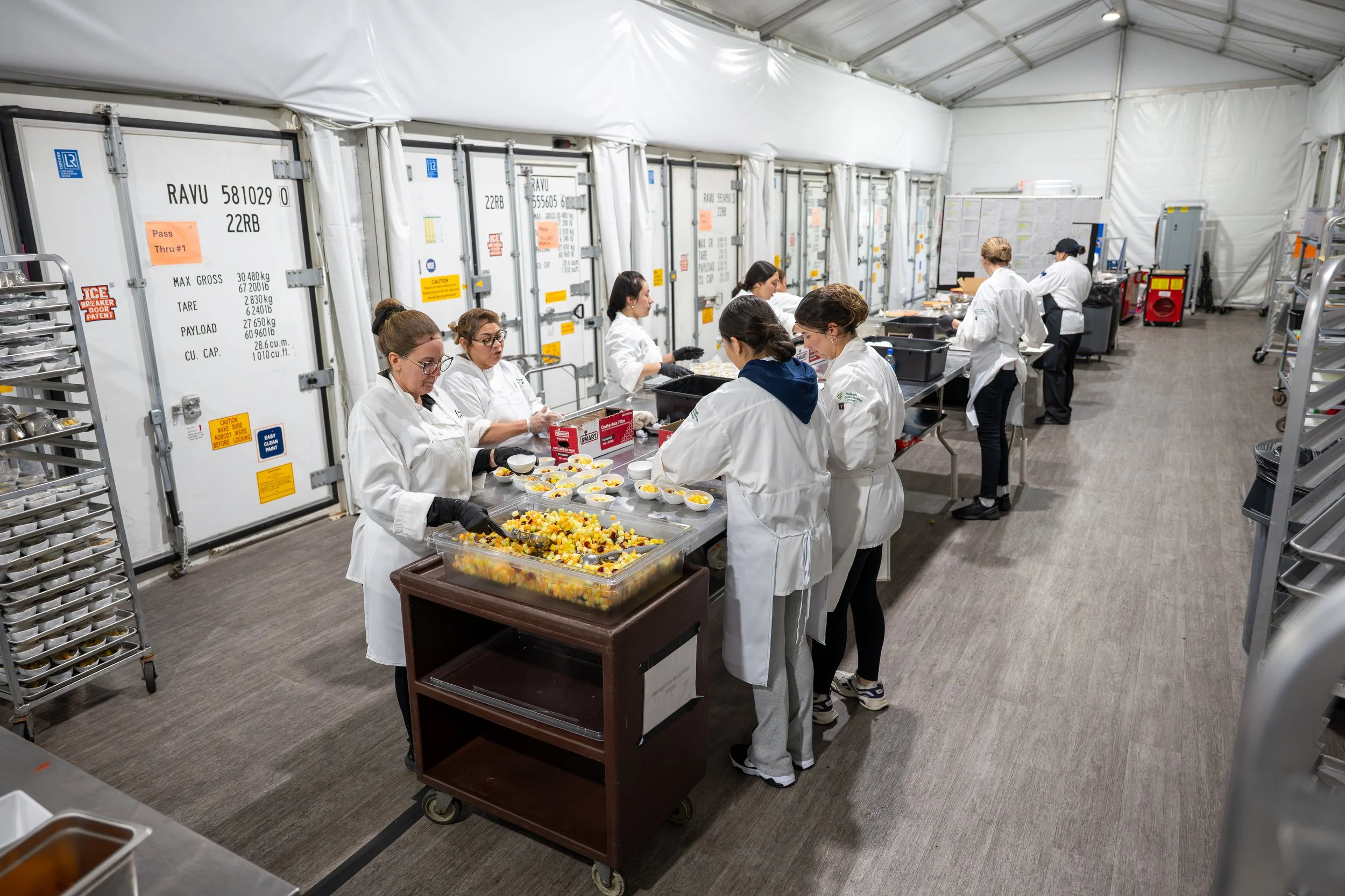 Workers in white chef coats preparing food in a temporary food production facility with large refrigeration units and shelves in the background.