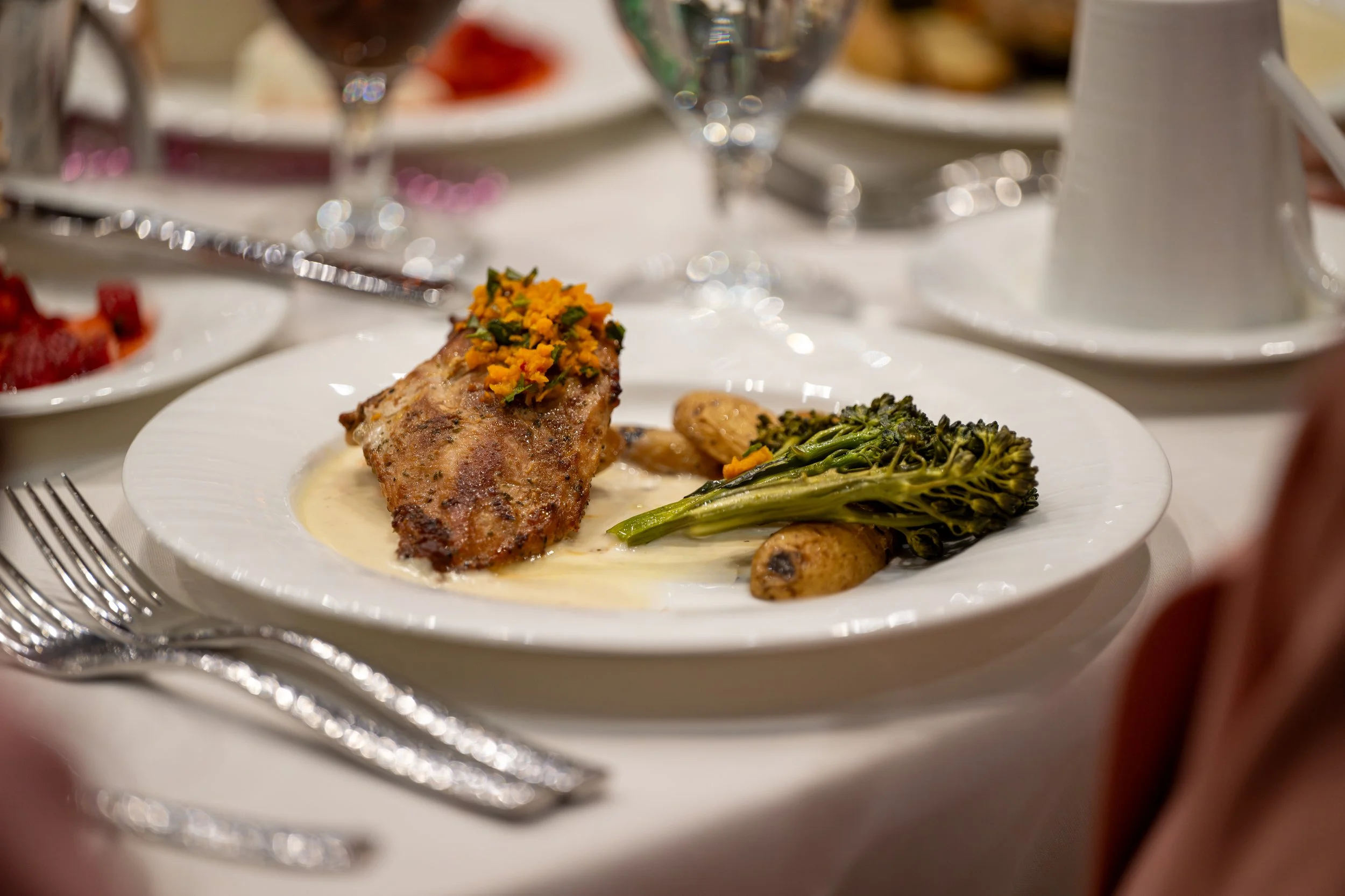 A plate of roasted chicken with herbs and vegetables including green broccolini and roasted potatoes, served on a white dinner plate at a formal dining setting.