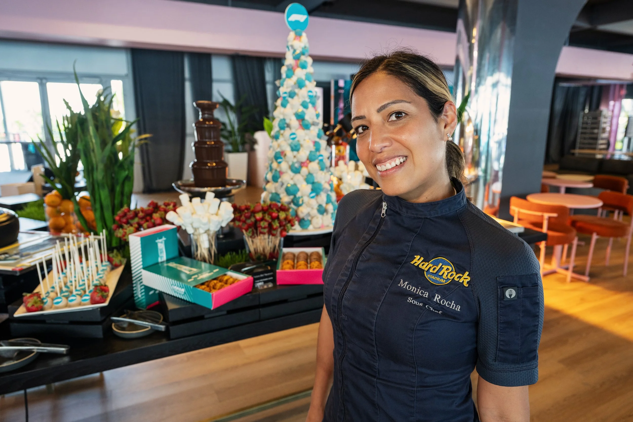A woman in a navy blue jacket with a Hard Rock logo and name tag standing next to a table with desserts and treats at a celebration event indoors, with a large layered dessert in the background.