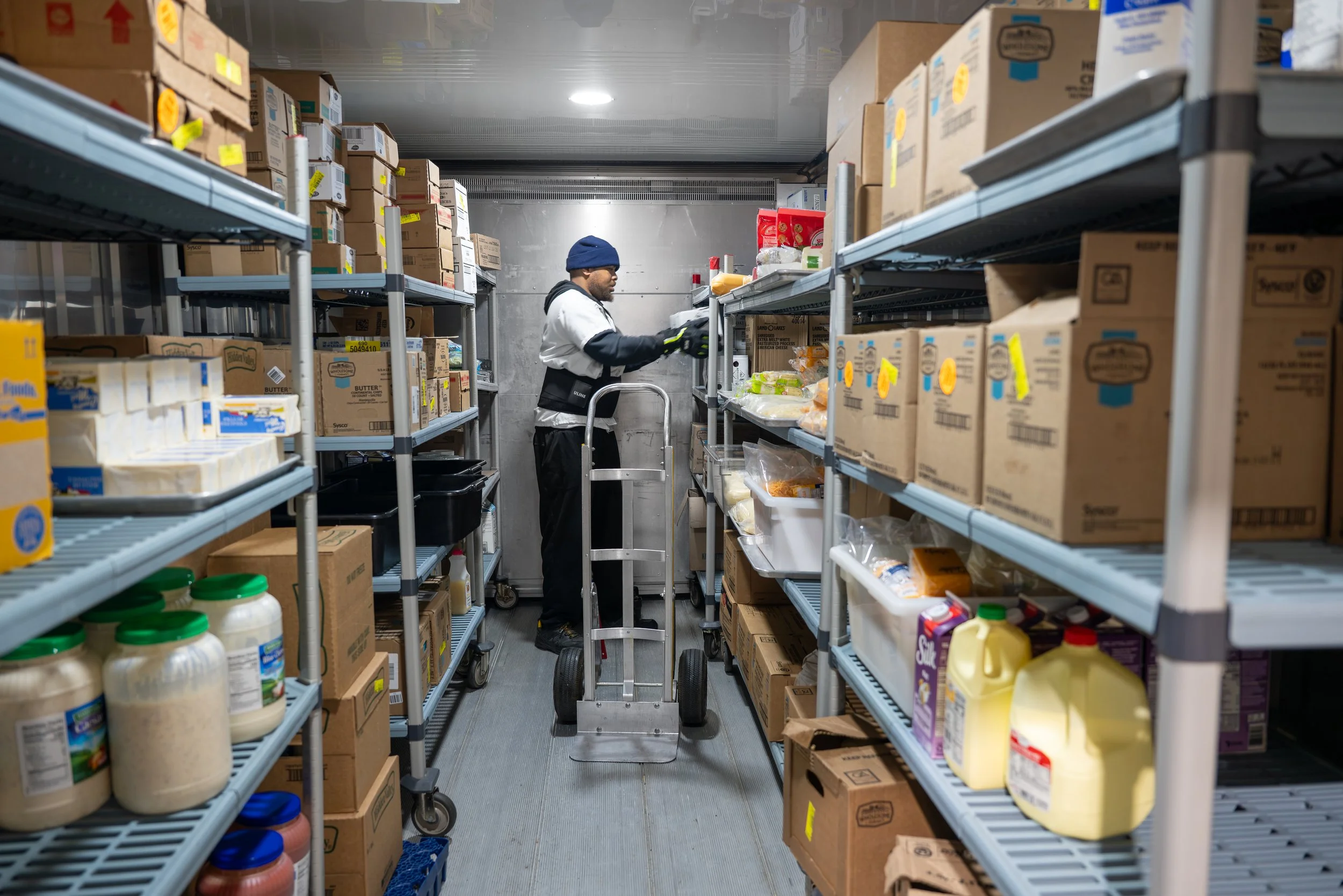 A person wearing a blue beanie, glasses, a white jacket with black accents, and gloves is standing on a small rolling step ladder in an industrial storage room, organizing or inspecting food products on metal shelves filled with boxes and bottles. Th