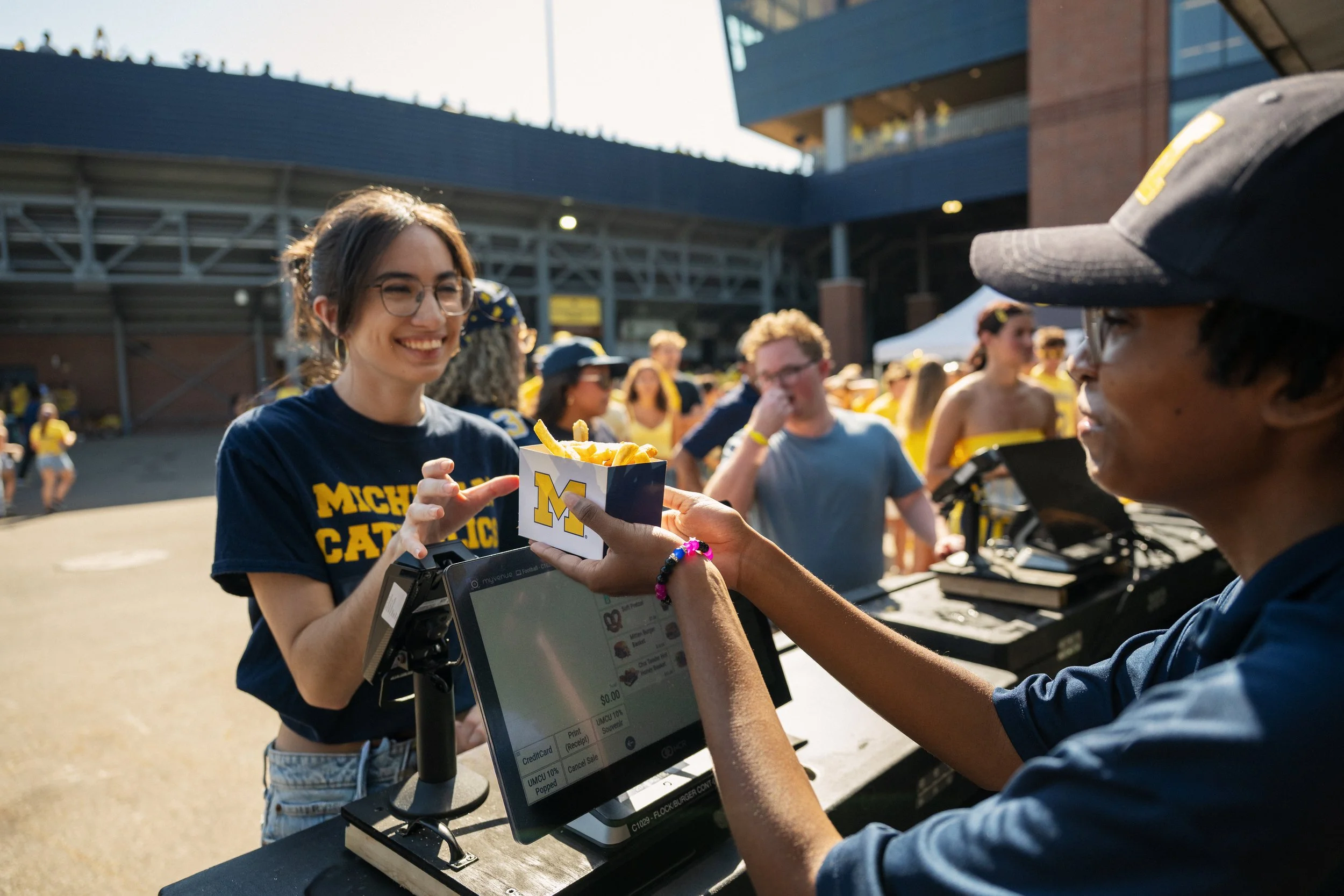 A young woman wearing glasses and a Michigan football shirt receives food from a man at an outdoor event, with several people in the background.