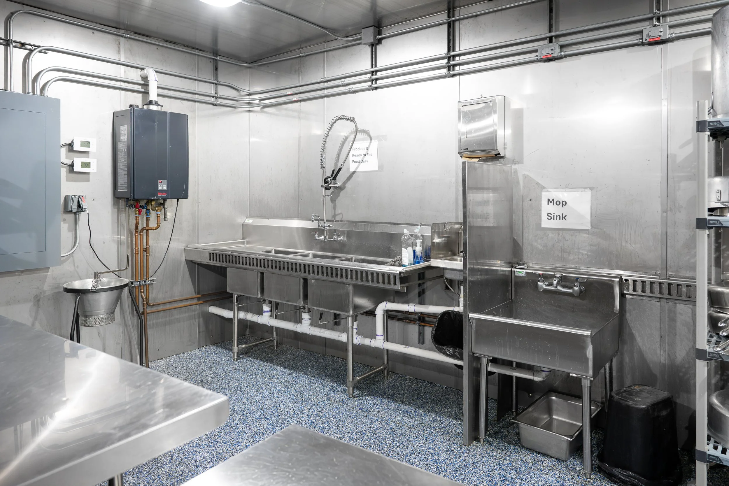 A commercial kitchen or food processing area with stainless steel sinks, cabinets, and equipment, including pipes and a water heater on the wall. There are cleaning supplies near the sink and signs on the wall indicating food safety.