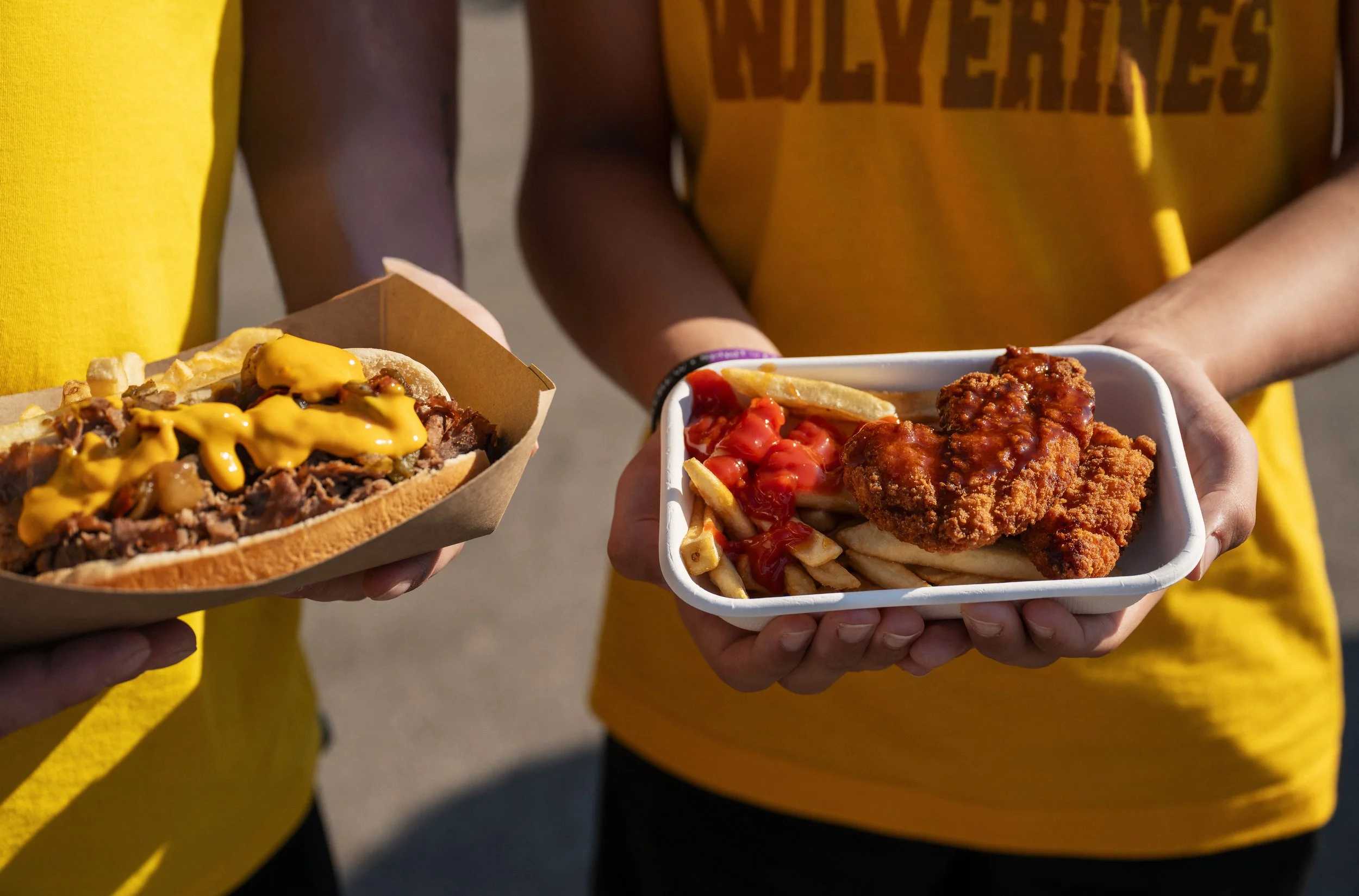 Two people holding food: one with a hot dog with mustard and toppings, the other with a tray of fries and fried chicken, both wearing yellow shirts.
