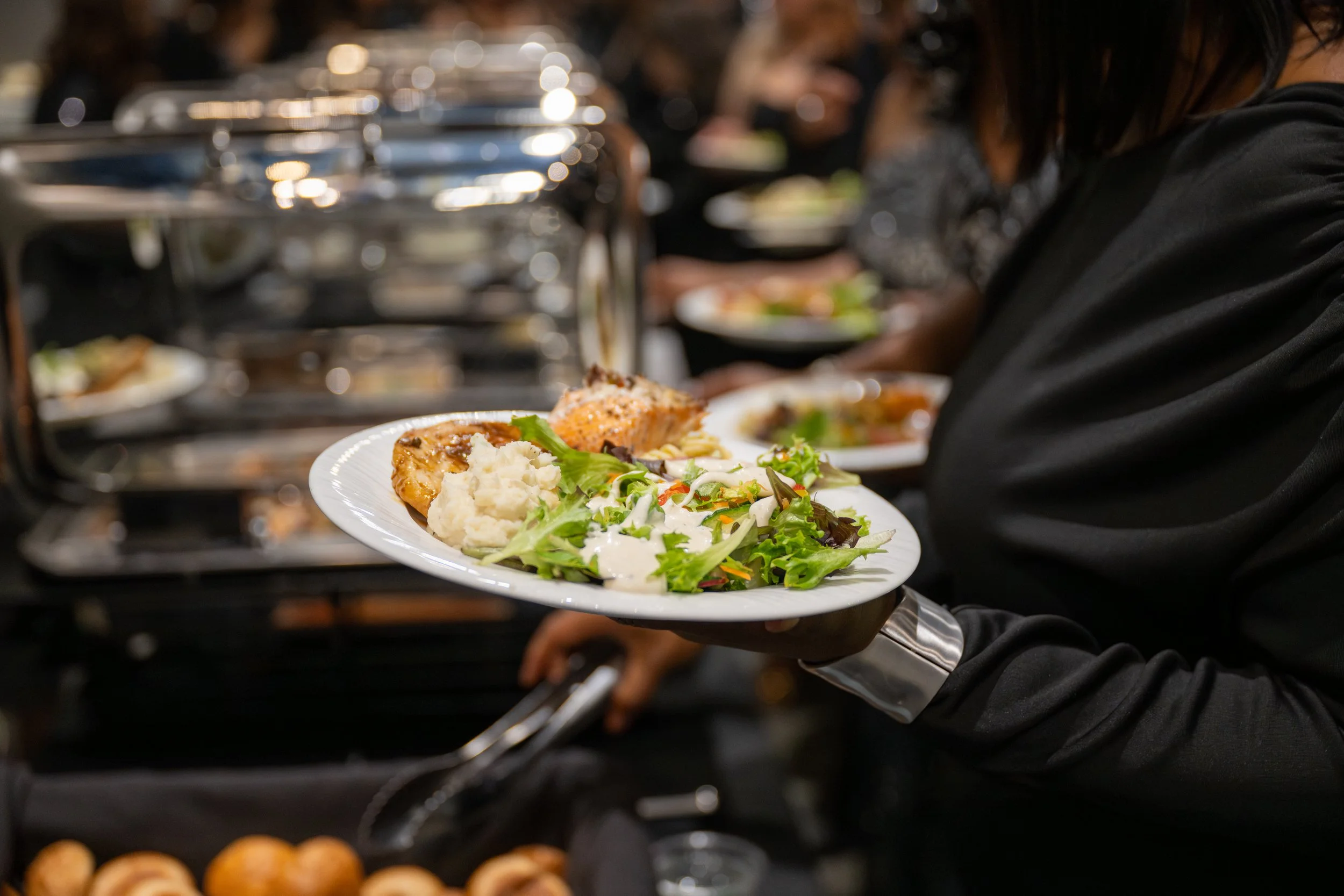 Person holding a plate of food with mashed potatoes, salad, and baked salmon at a buffet line.