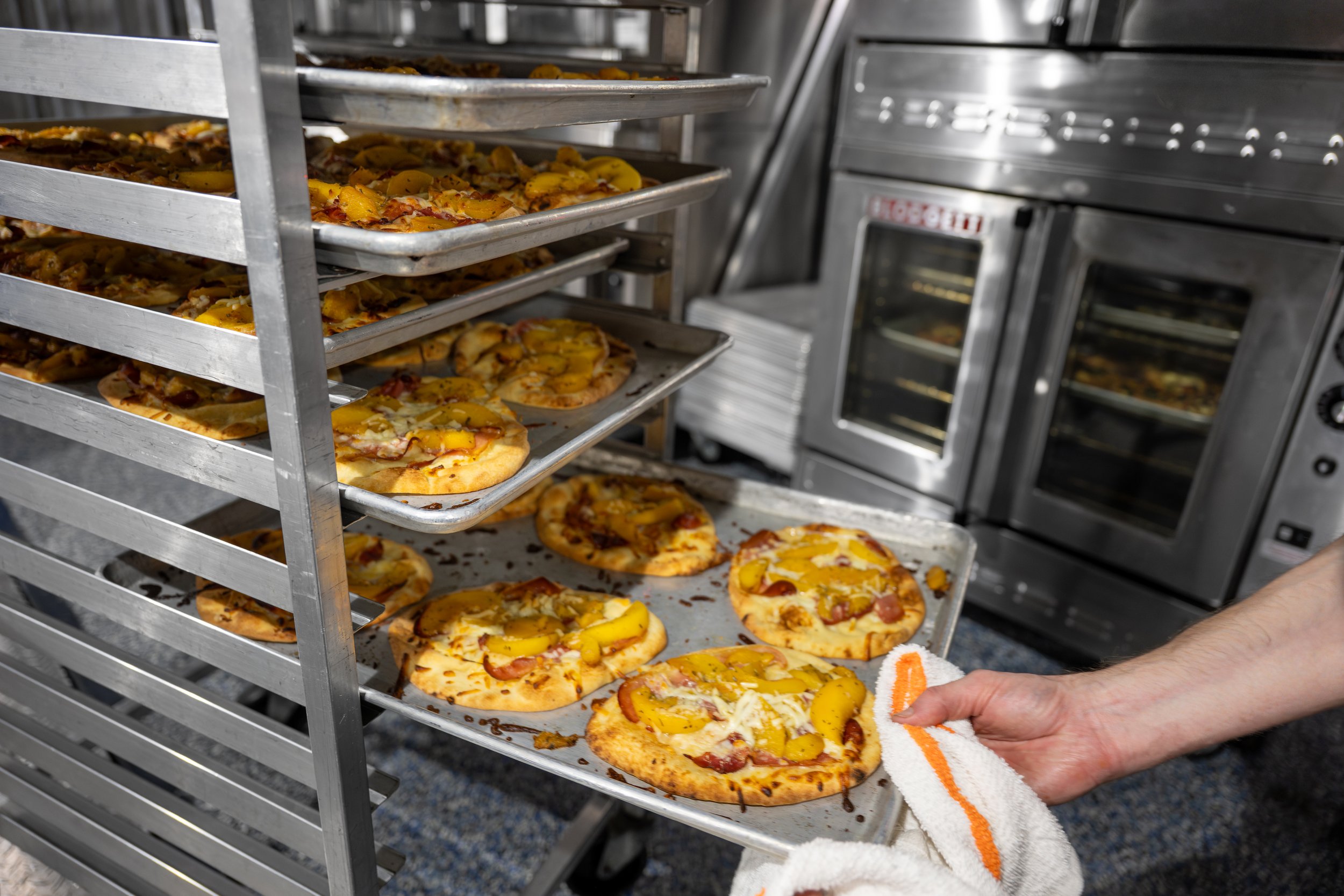 A person is removing a tray of small pizzas topped with cheese, sliced peaches, and meats from an industrial oven in a commercial kitchen.