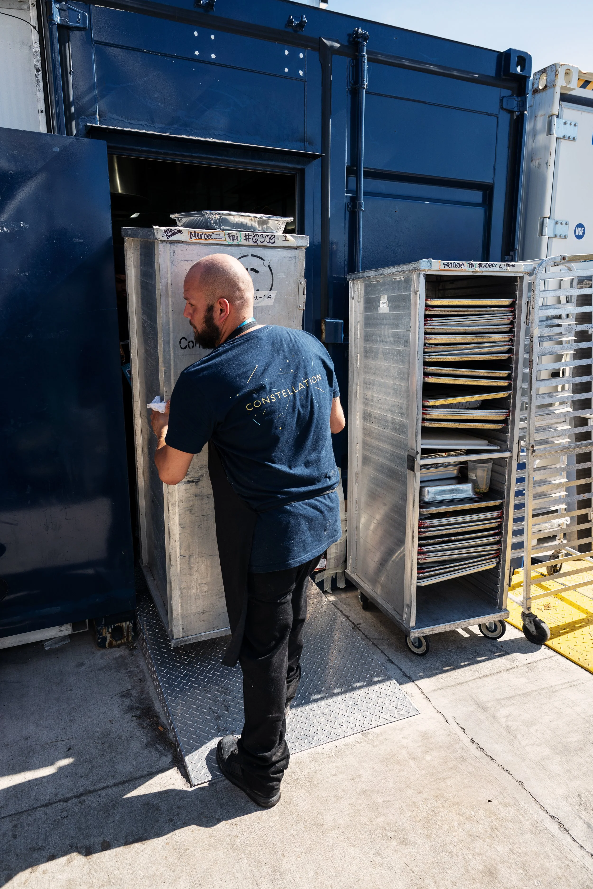 A man with a shaved head and beard, wearing a black t-shirt with the word 'CONSTELLATION' on the back, is cleaning a metal door or cabinet with a cloth outside a shipping container. There are metal racks with trays or pans nearby, and a cart with sta