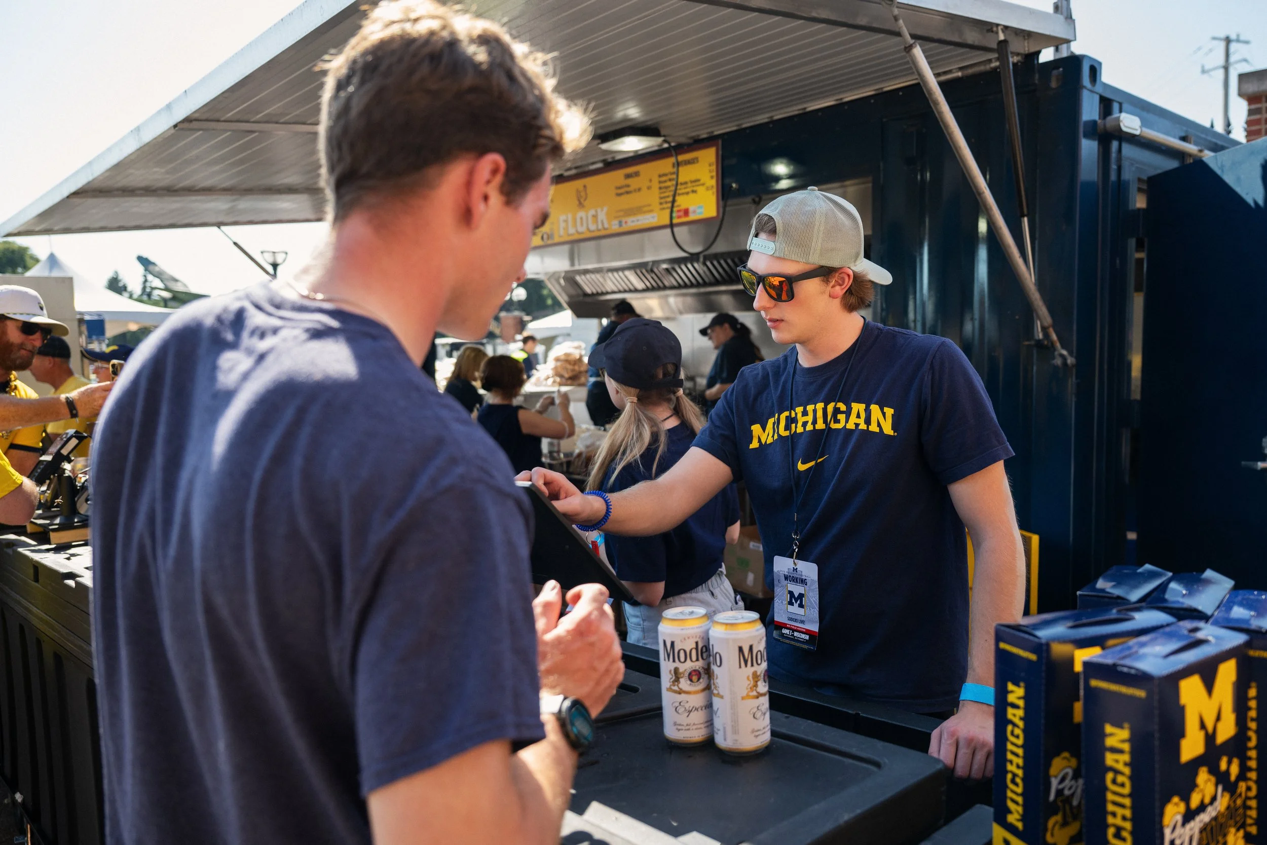 A young man wearing sunglasses and a Michigan shirt at a food stall, with aluminum cans of Modelo beer in the foreground.