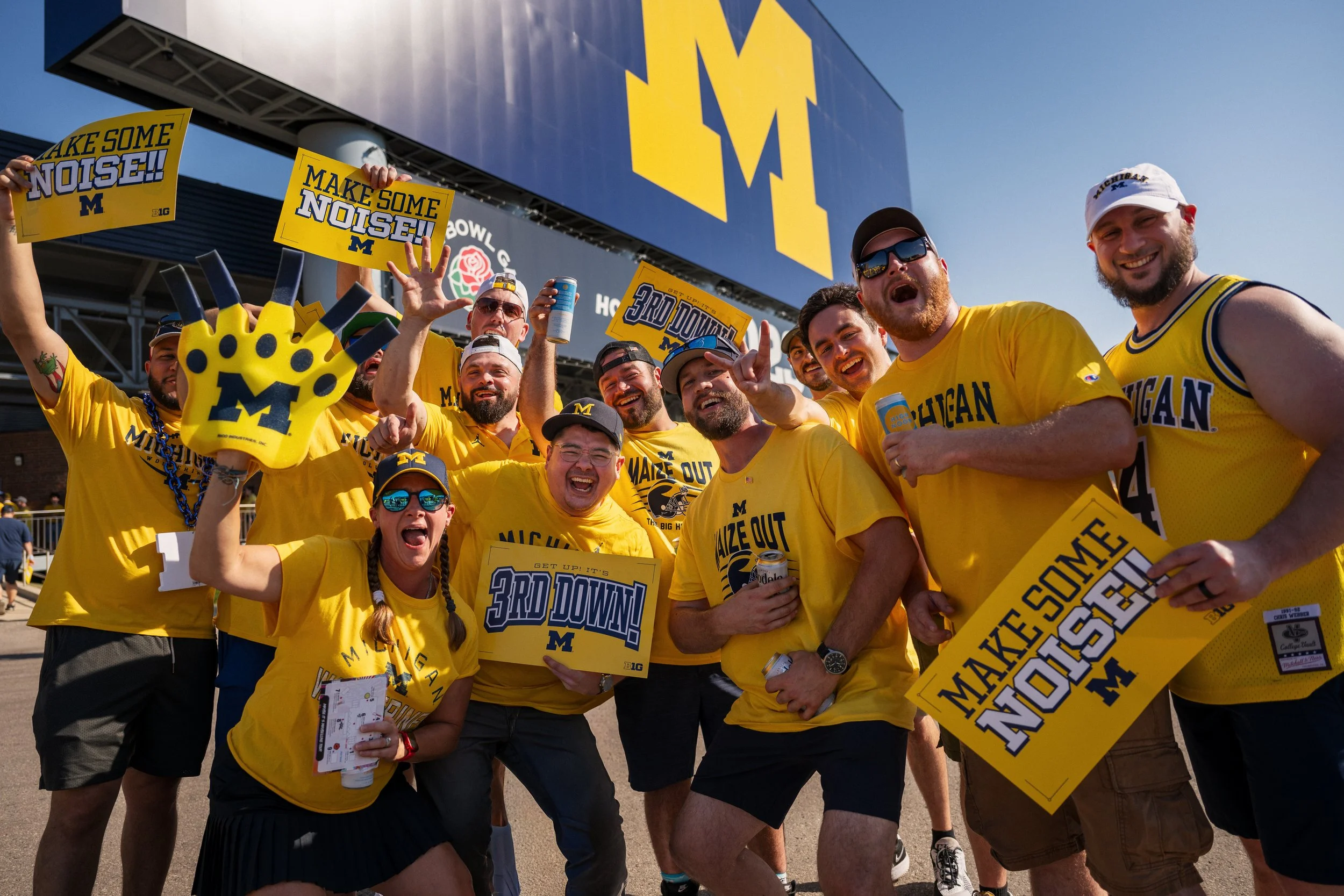 Group of University of Michigan football fans celebrating outside the stadium in yellow shirts, holding signs that say 'Make Some Noise!' and '3rd Down!', with a large Michigan logo billboard in the background.
