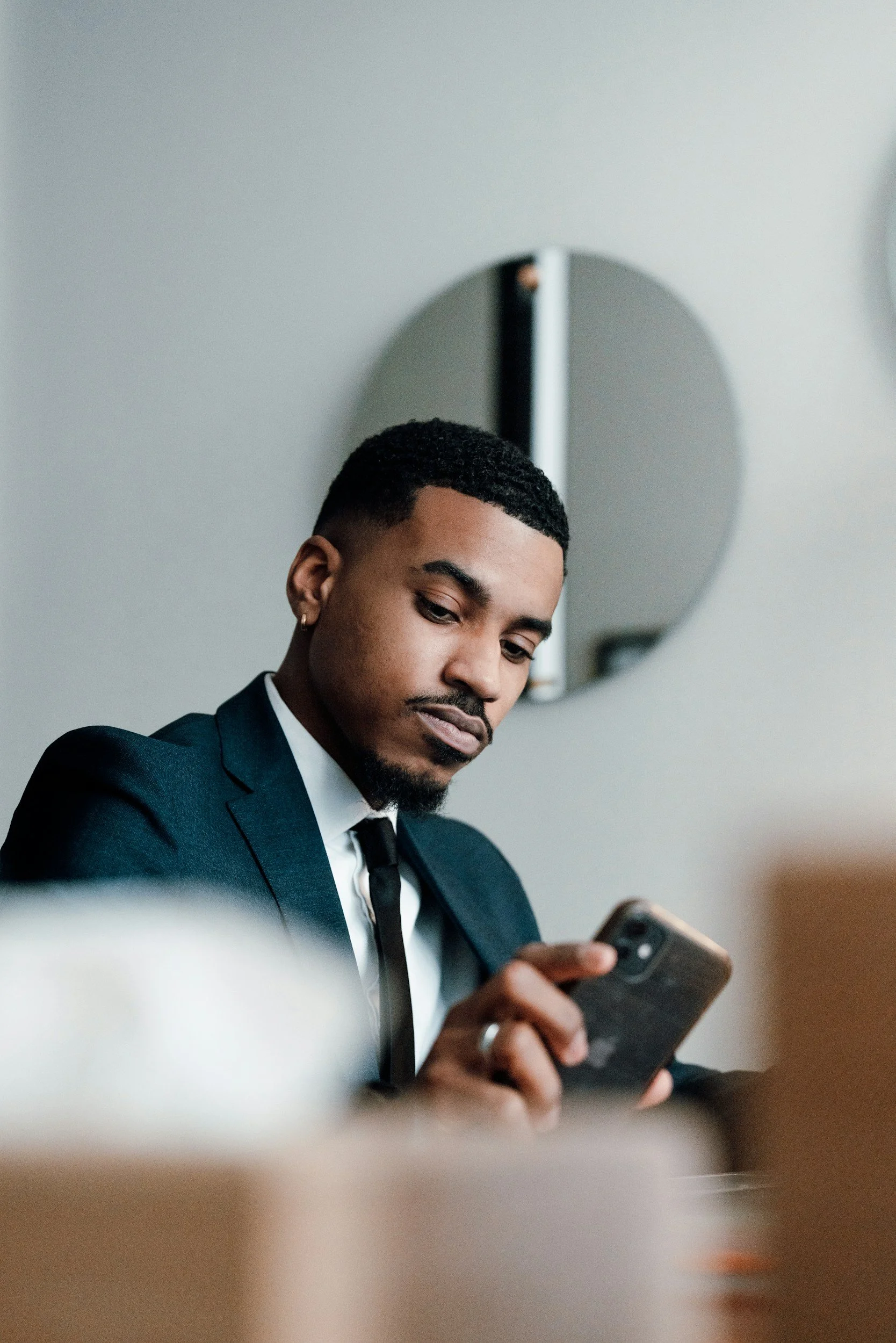 A young man in a dark suit with a white shirt and black tie, with short black hair and a beard, looking at a smartphone while seated in a modern room with a circular mirror on the wall behind him.