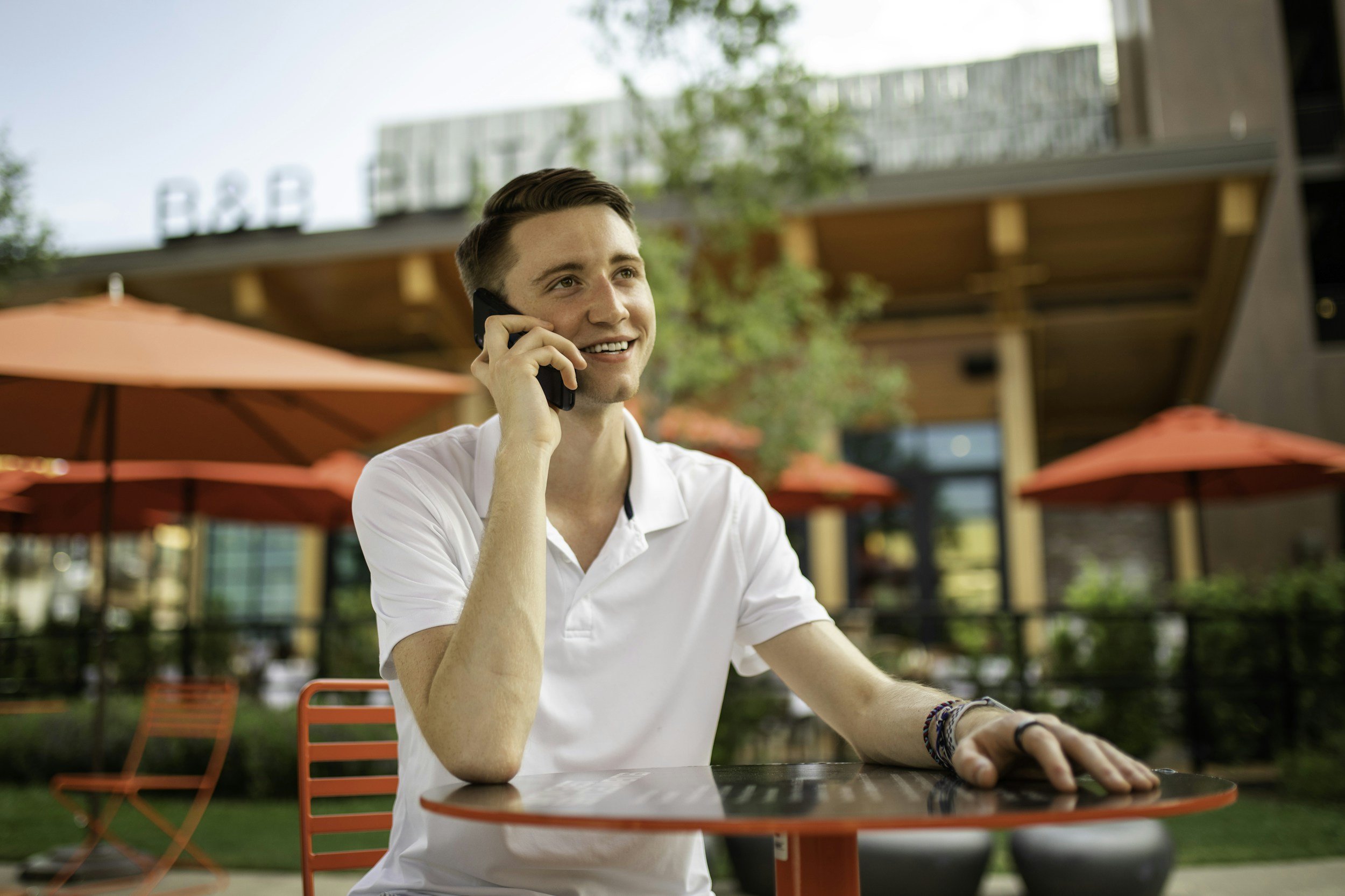 Young man smiling and talking on a cell phone while sitting at an outdoor cafe table with red chairs and red umbrella, with a modern building in the background.