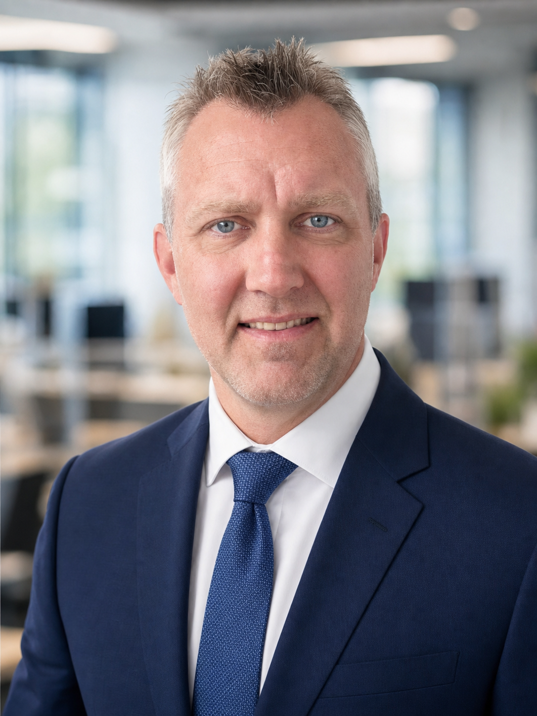 Headshot of a middle-aged man with short, spiky gray hair, blue eyes, and fair skin, wearing a navy suit, white dress shirt, and blue tie, in an office setting with blurred background windows and desks.