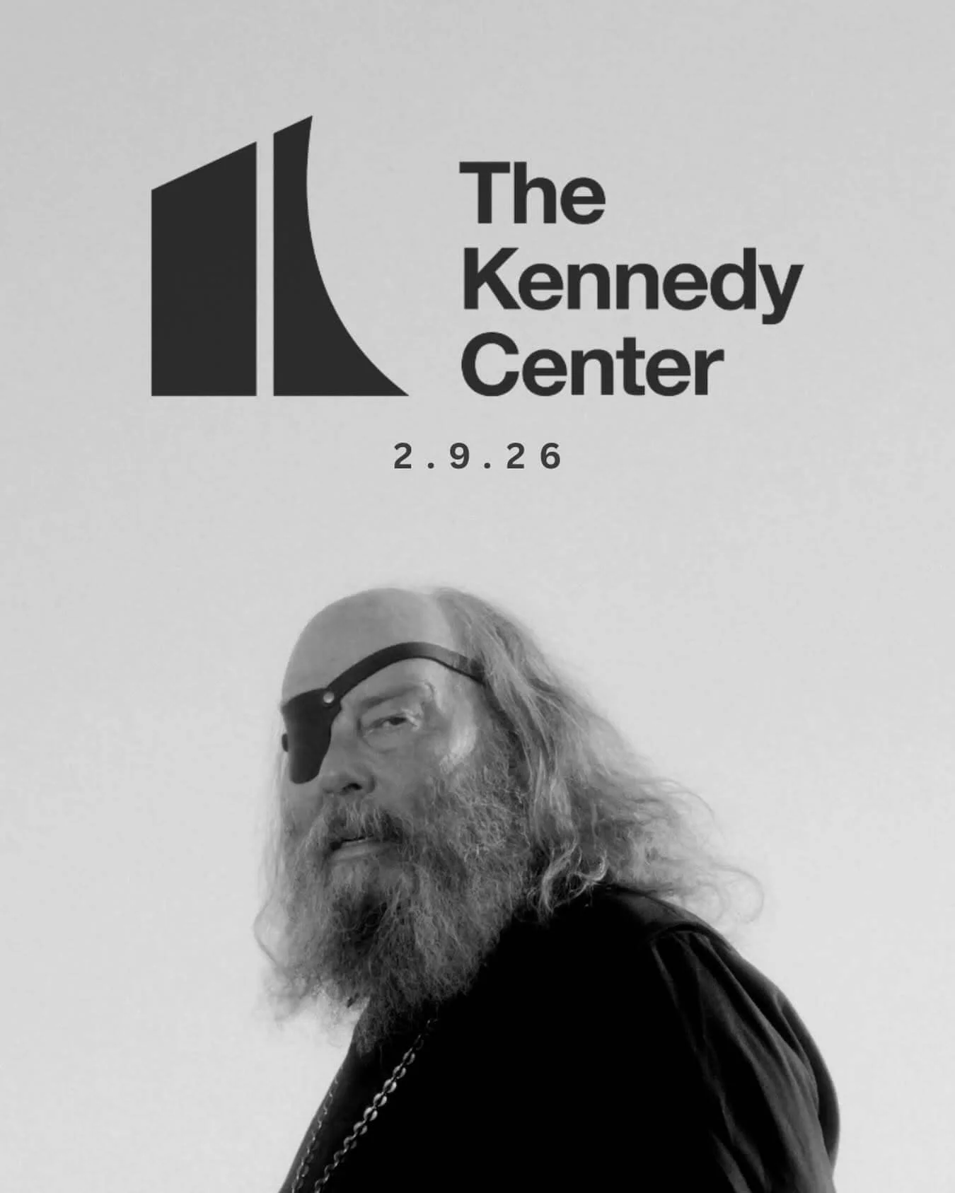 A man with long gray hair, a beard, and wearing dark sunglasses, standing in front of a wall with the logo and text for The Kennedy Center and the date 2.9.26.