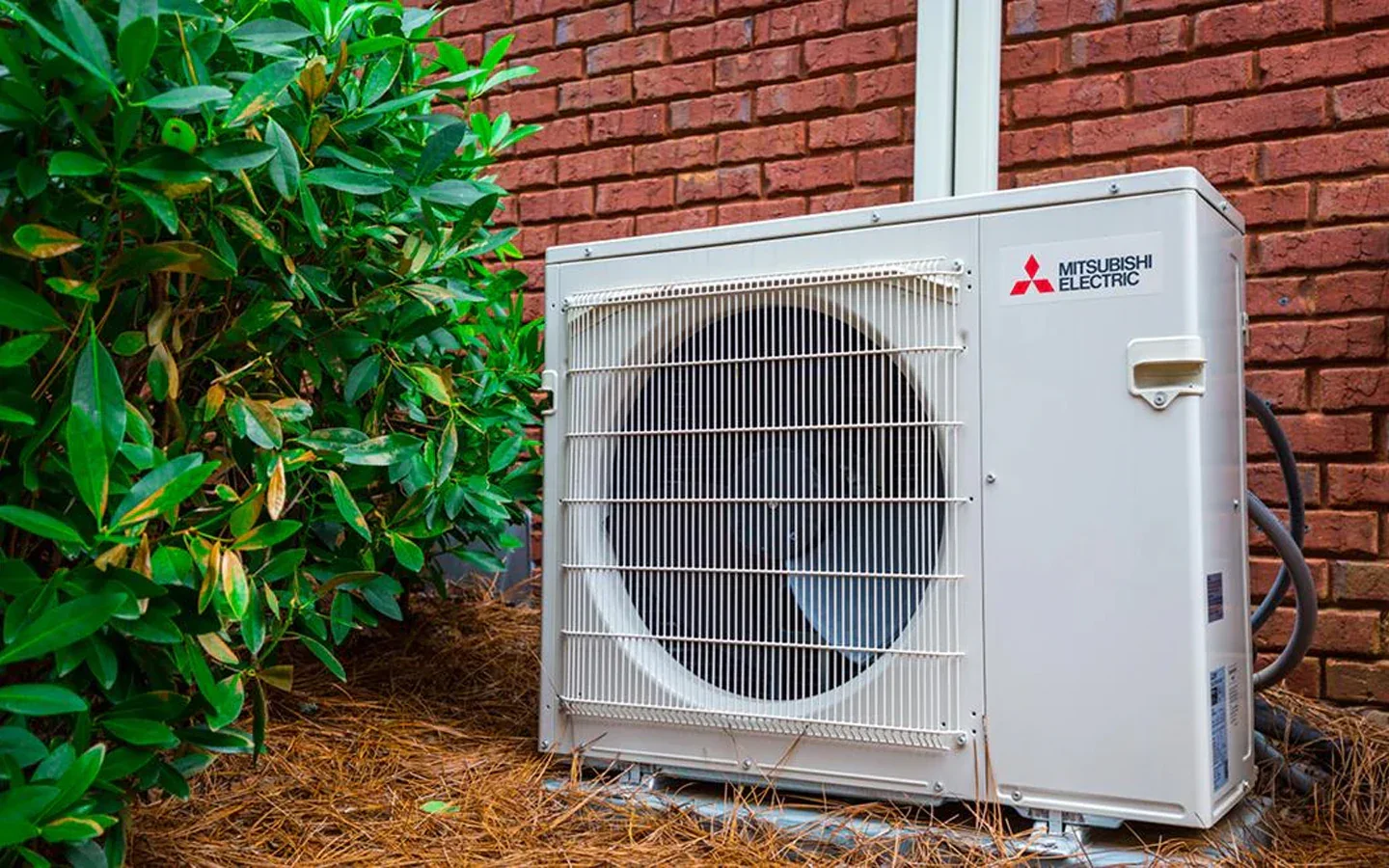 Outdoor Mitsubishi Electric air conditioning unit next to a brick wall and green bushes with pine straw ground cover.