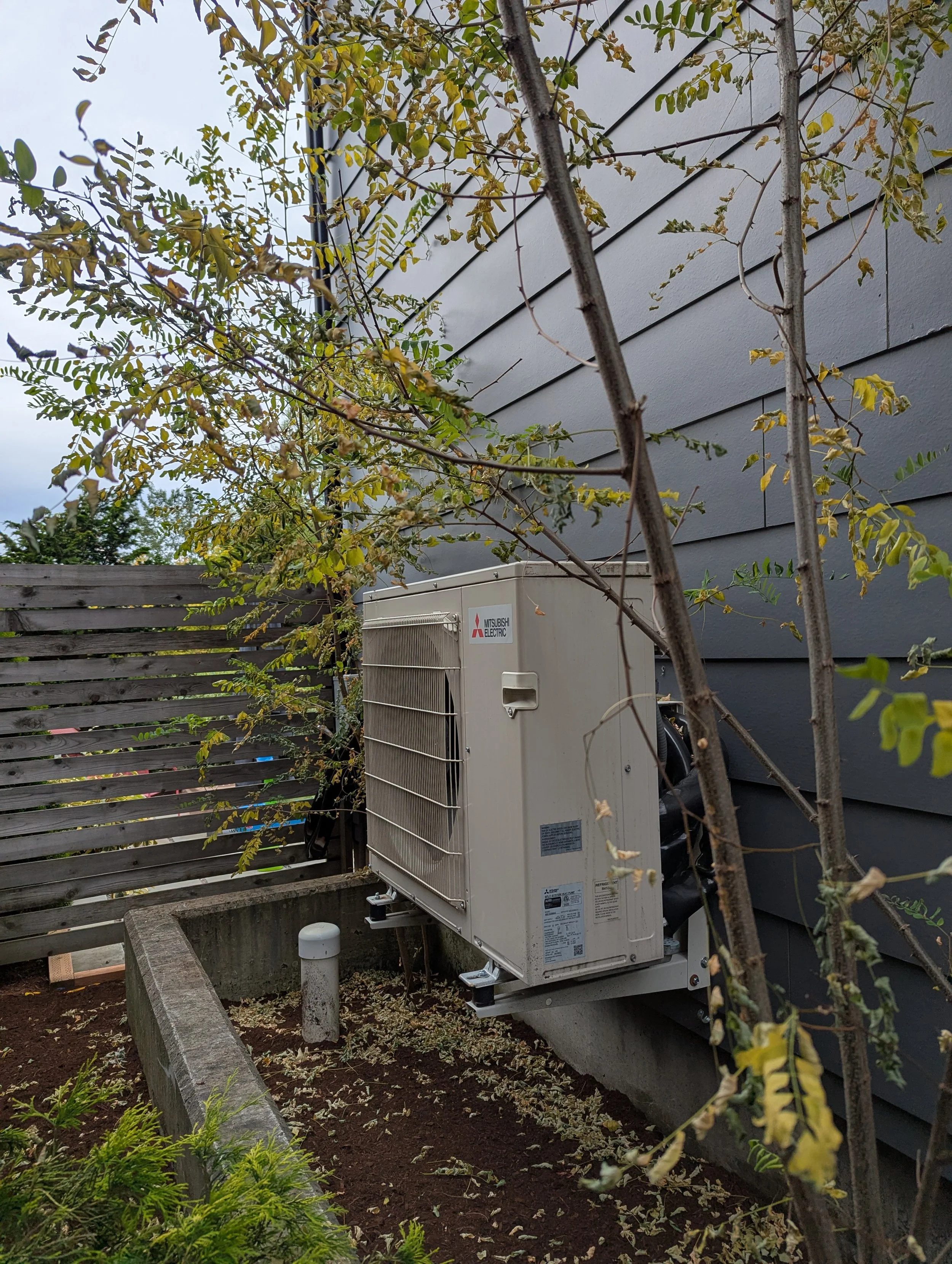 Mitsubishi condenser unit, outdoors resting on a wall mount, surrounded by bamboo trees and garden plants.