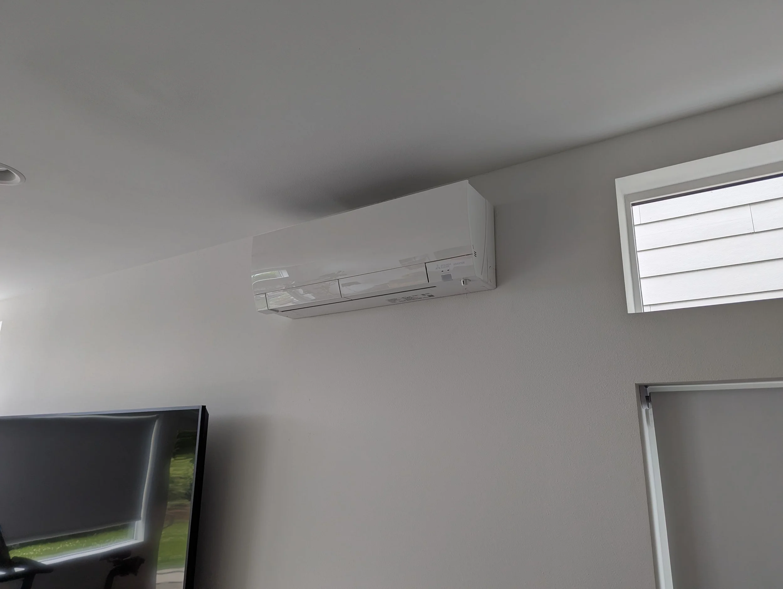 White wall with a wall-mounted air conditioning unit and a window with horizontal blinds, part of a living room or office space.