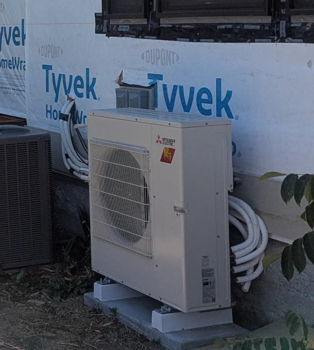Mitsubishi condenser unit, outdoors on a pedestal resting on the ground next to a home, with Tyvek weather barrier on the building wall.