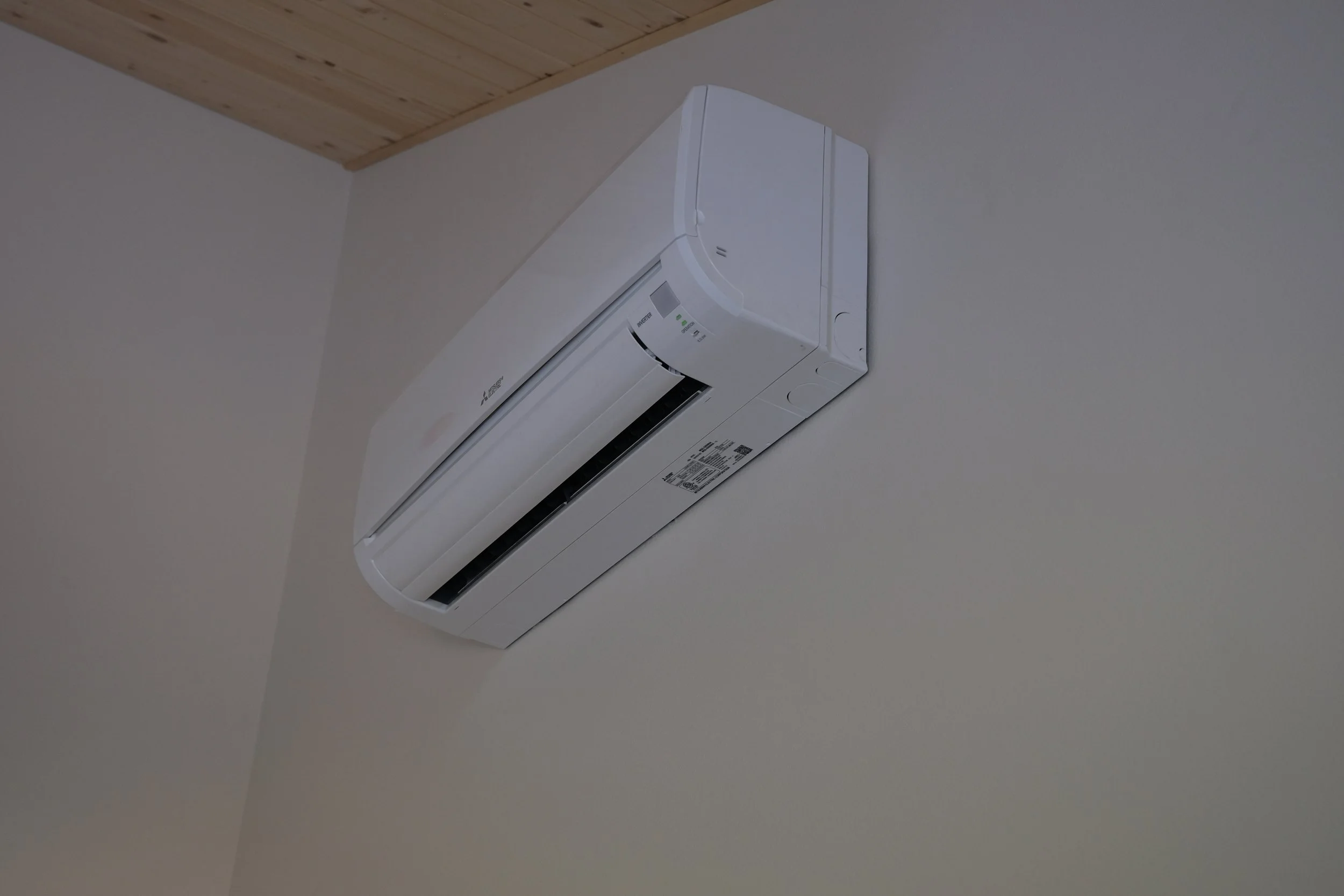 Wall-mounted white Mitsubishi air conditioning unit installed near the ceiling on a plain wall, with a wooden ceiling visible at the top of the image.