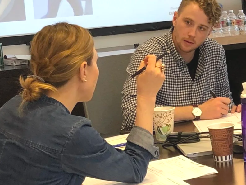 Two people engaged in a discussion at a meeting or seminar, with papers, notebooks, and coffee cups on the table.