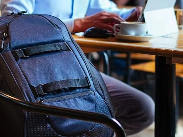 Close-up of a blue backpack on a chair in a café, with a person in the background working on a laptop at a wooden table, drinking coffee.