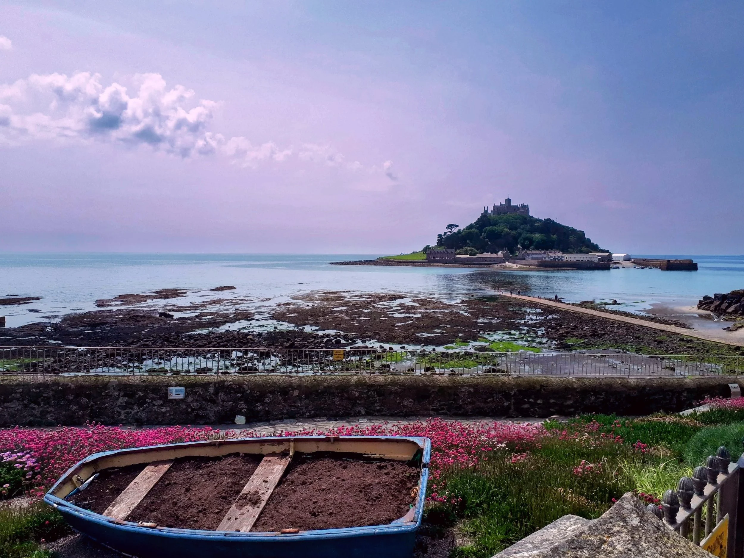 A coastal scene at low tide, with a traditional boat in the foreground and St Michael's Mount in the distance. Captured in soft natural light, offering a tranquil view of the Cornish coastline.