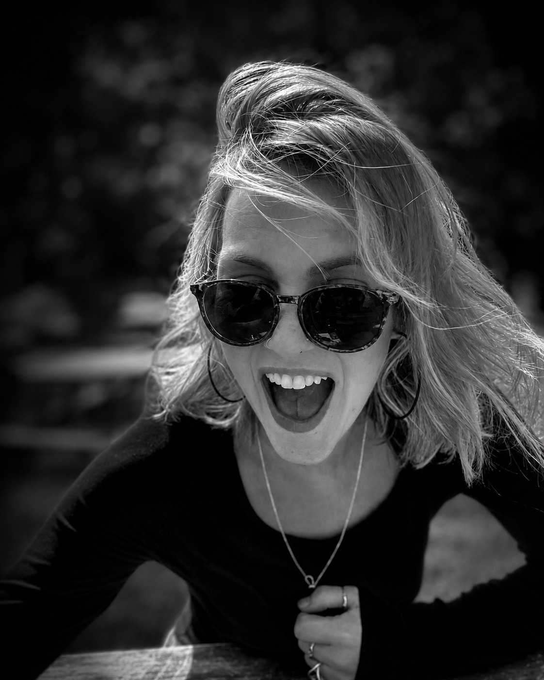 A woman with blonde hair, wearing round sunglasses, laughing with her mouth open, outdoors with blurred background, in black and white.