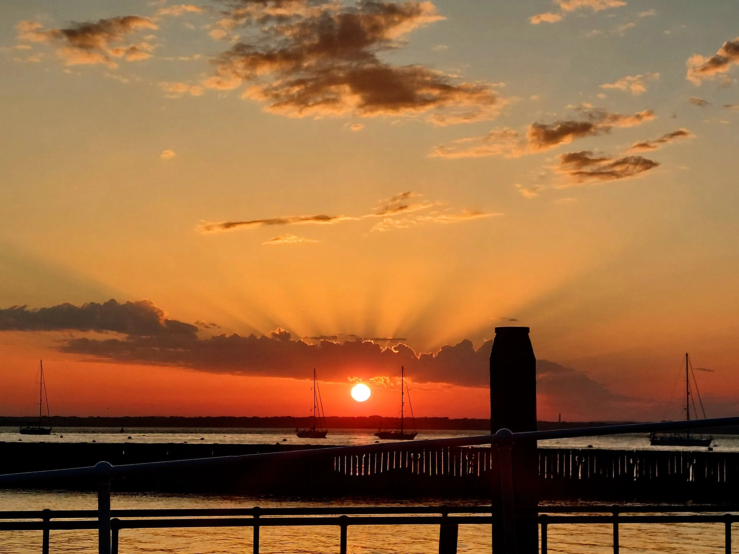 The combination of glowing sunset colors, radiant sunbeams, still water, and silhouetted boats creates a tranquil coastal scene often associated with the end of a summer day.