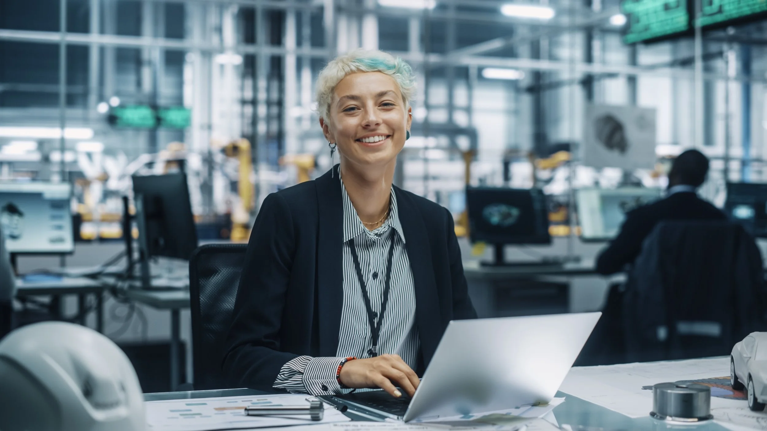 A smiling woman with short, platinum blonde hair, sitting at a desk in an industrial office environment, working with a laptop and papers.